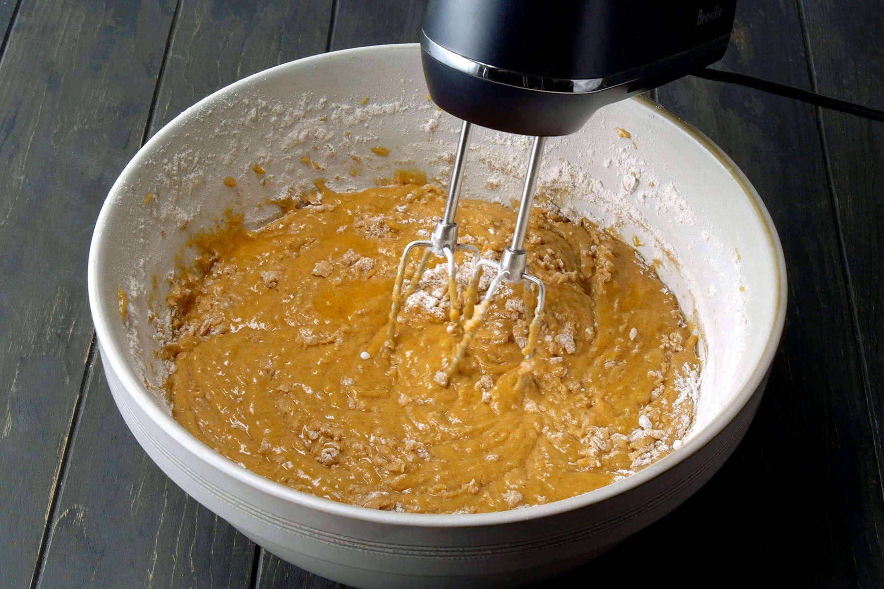 A black electric handheld mixer blending brown dough in a large white mixing bowl on a dark wooden surface. Flour is scattered around the sides of the bowl.