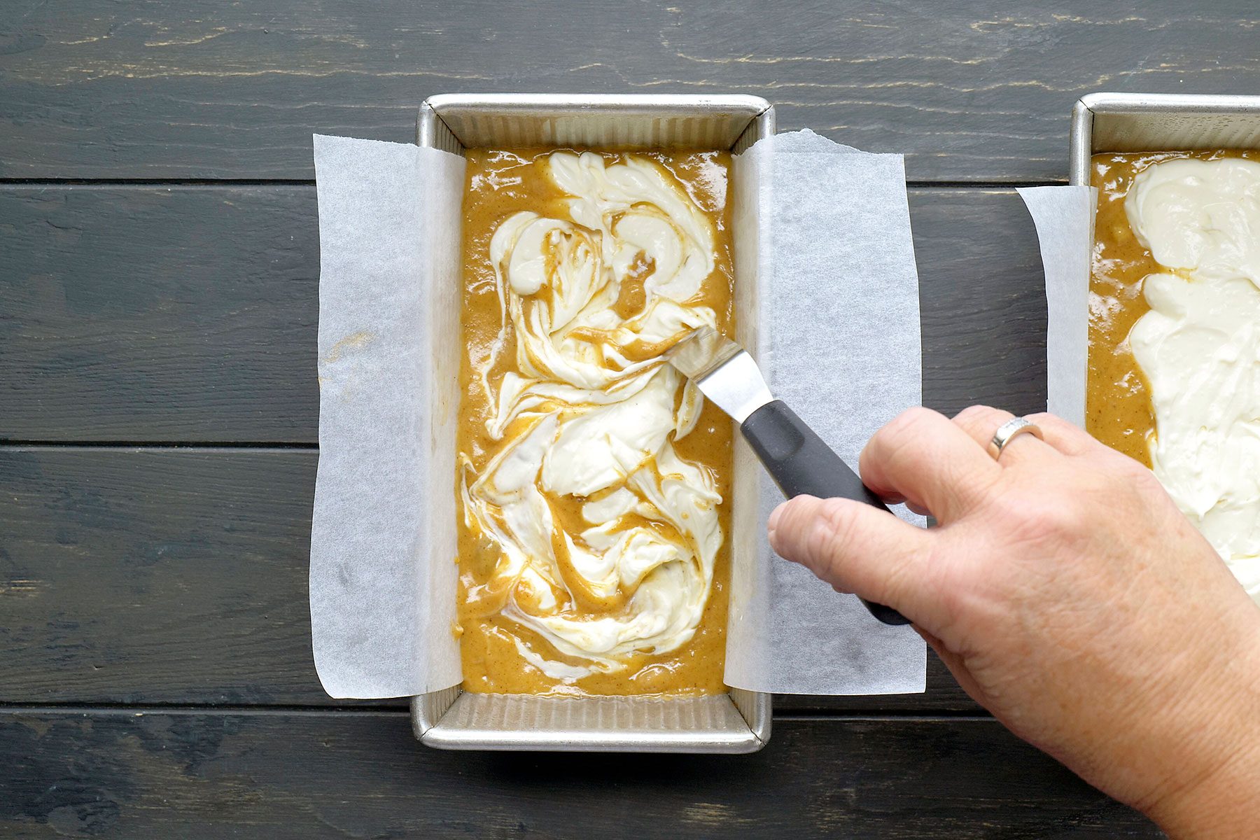 A person spreads cream cheese mixture into pumpkin batter in a rectangular baking pan lined with parchment paper. The mixing creates a swirl pattern. A metal spatula with a black handle is used to blend the layers together.