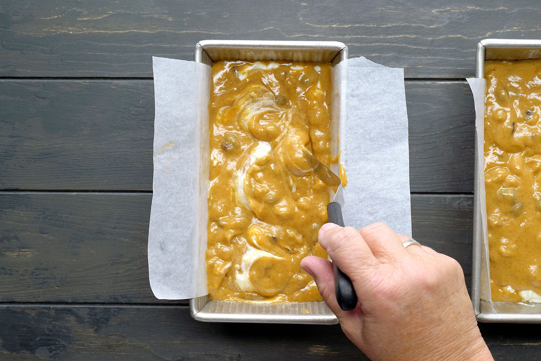 A hand spreads banana bread batter in a rectangular baking pan lined with parchment paper. The batter appears lumpy with visible chunks, and the pan rests on a dark wooden surface.