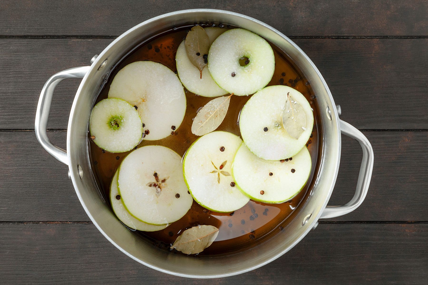A pot on a dark wooden surface containing sliced green apples, bay leaves, and peppercorns in a liquid, likely being prepared for cooking or flavoring.
