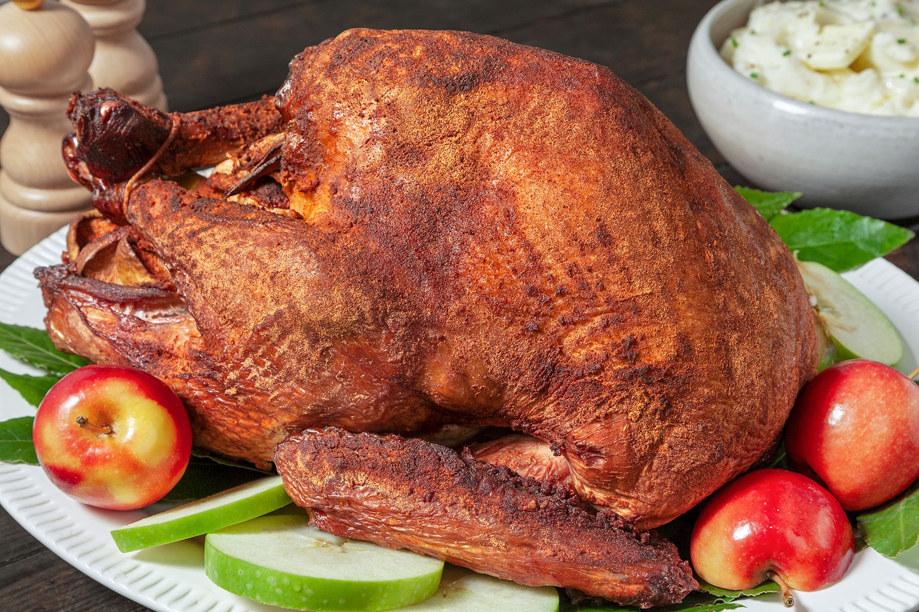 A roasted turkey on a white platter is garnished with green apple slices and red apples. In the background, there's a bowl of mashed potatoes. The turkey's skin is golden brown and crispy.