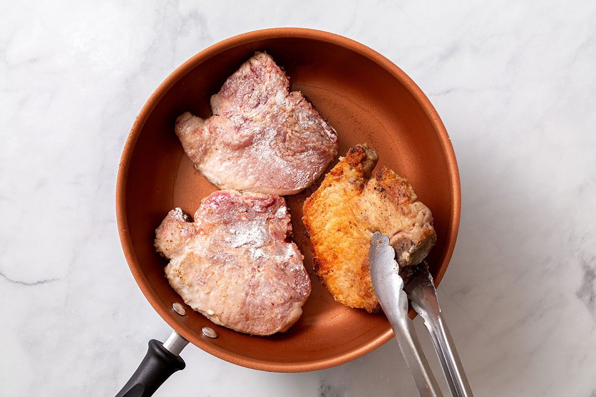 Flour coated pork chops being fried in a large skillet until browned for step two of Smothered Pork Chops in Oven recipe for Taste of Home