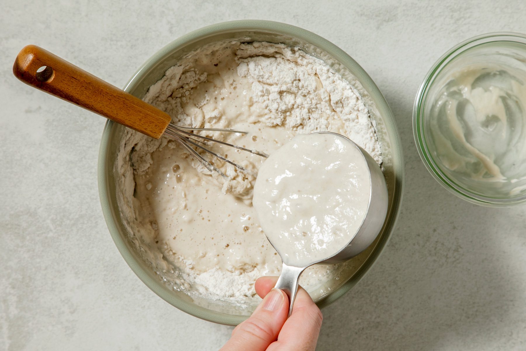 overhead shot of a person is pouring a liquid from a measuring cup into a bowl containing a dry mixture, The dry mixture appears to be flour, and the liquid is creating a well in the center of the mixture, A whisk is resting in the bowl;