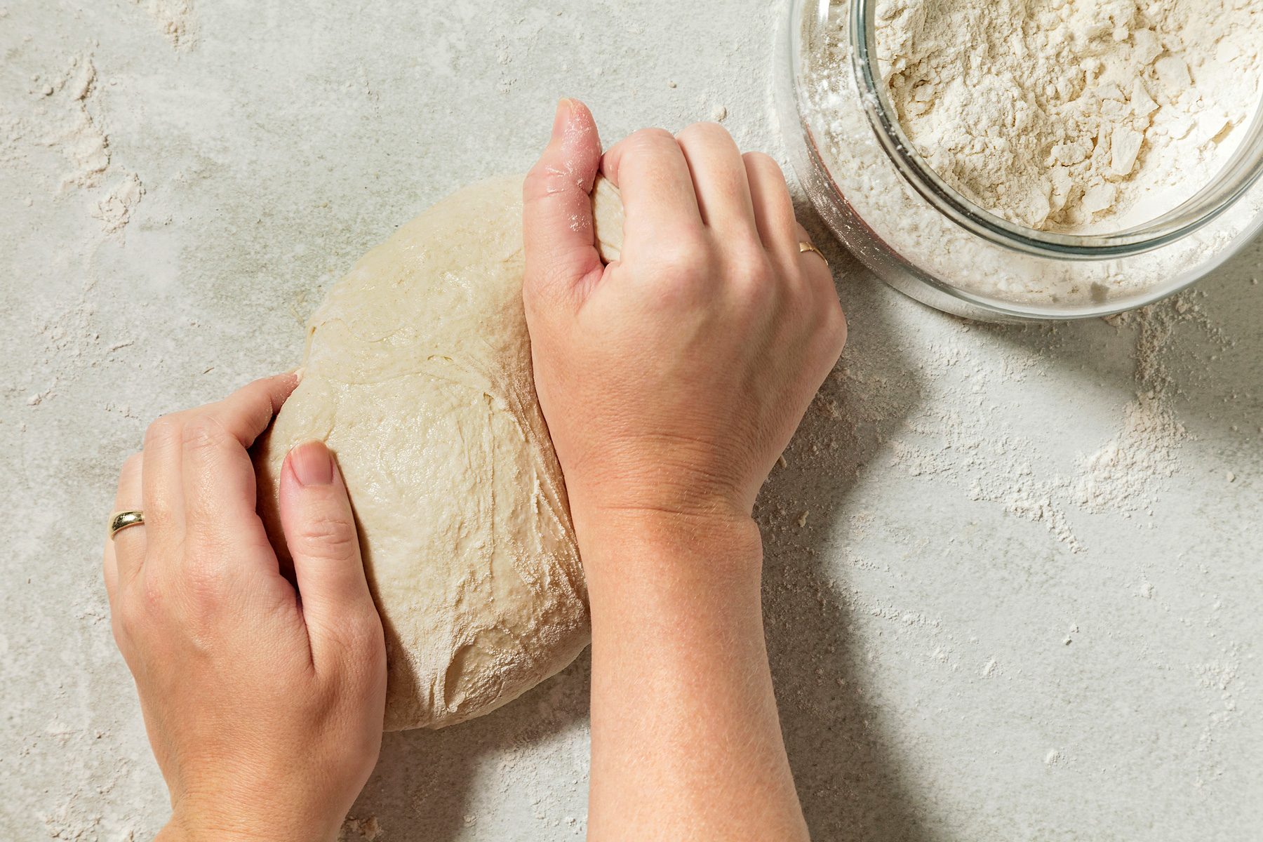 overhead shot of a pair of hands kneading a dough on a gray surface, The hands are pressing and folding the dough, working it into a smooth and elastic consistency, A jar of flour is visible in the background;