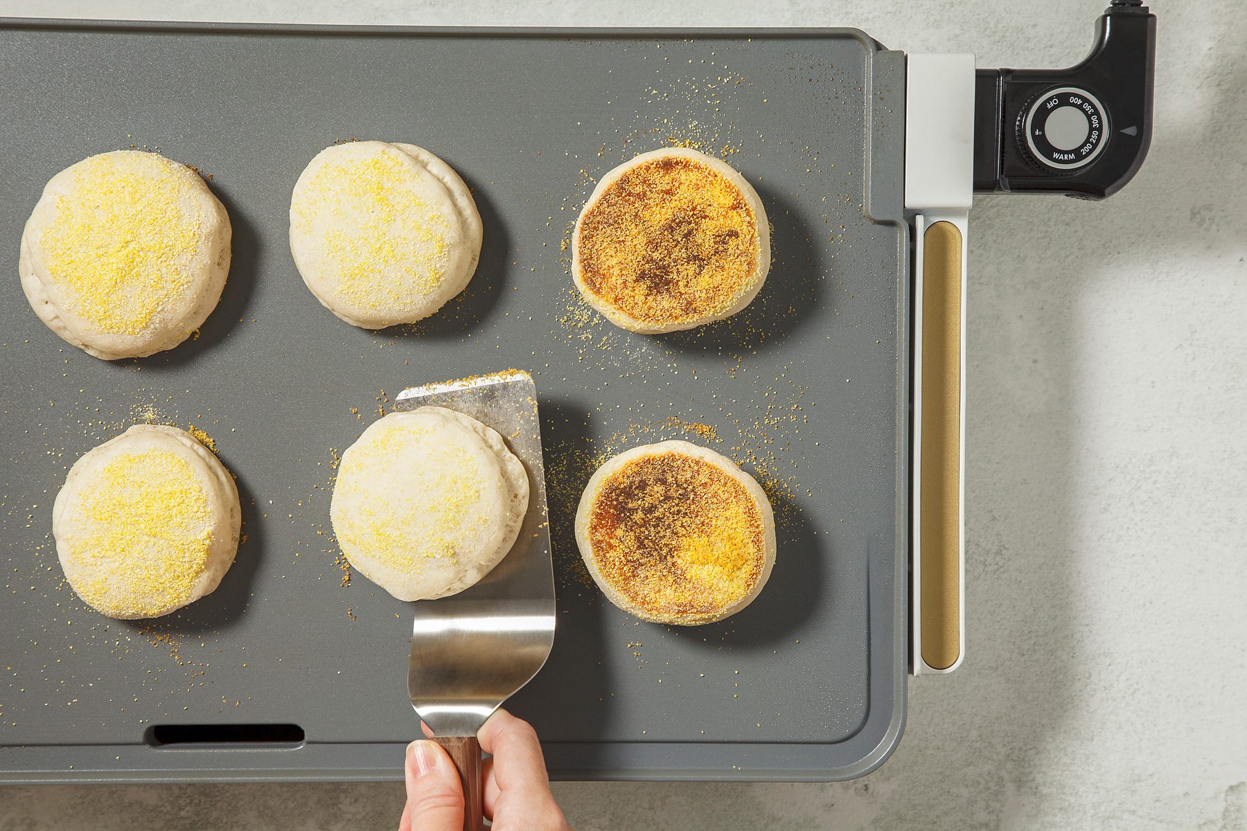 overhead shot of a griddle with several muffins cooking, The muffins are golden brown and are covered in a layer of cornmeal, A spatula is being used to flip one of the muffins;