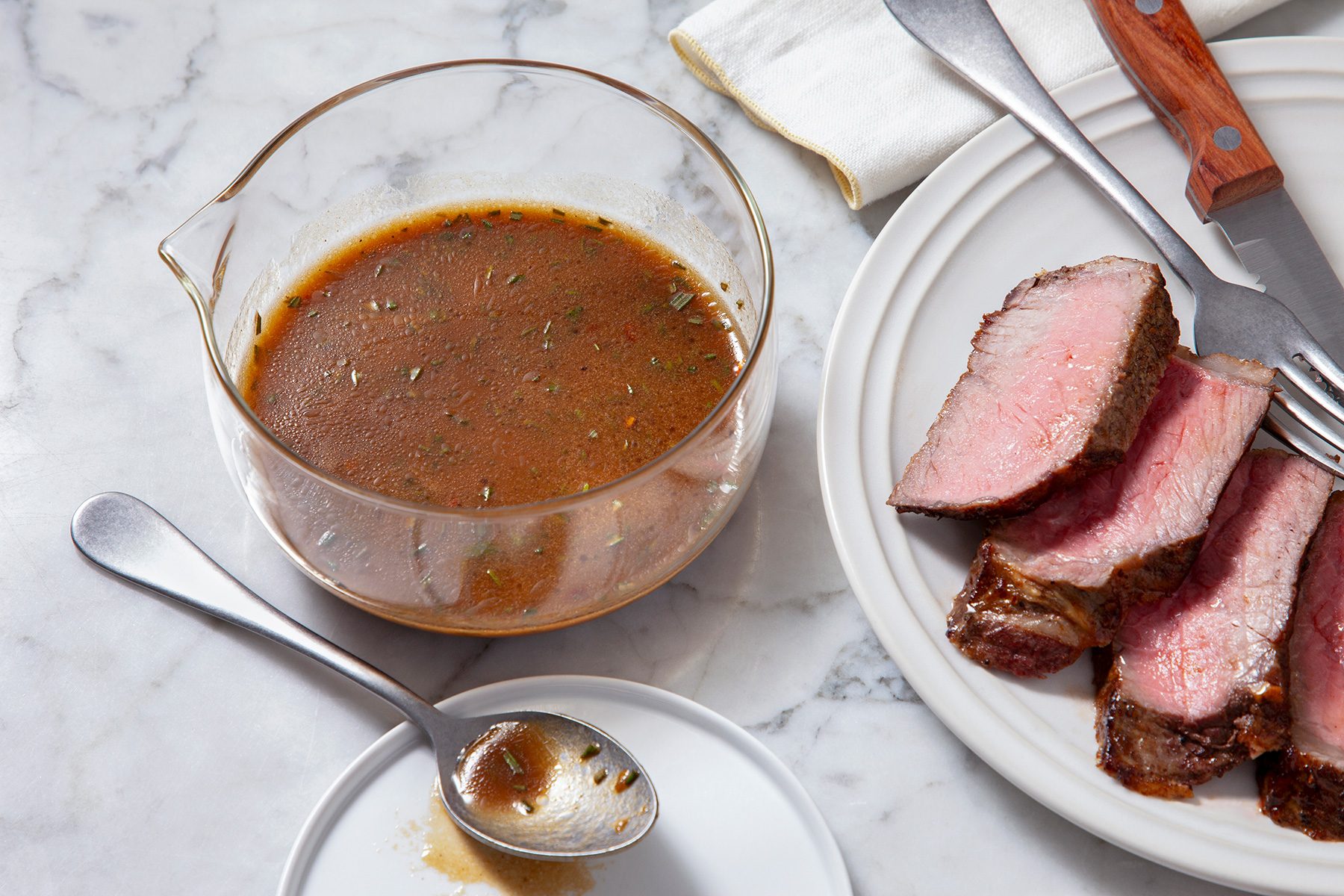 3/4th shot of a marinade and the meat, The focal point is a glass bowl filled with a rich, brown sauce or marinade, flecked with herbs and spices, To the right, a white plate holds slices of cooked meat, possibly beef, A carving fork and knife rest on the plate, In the foreground, a spoon rests on a small plate;