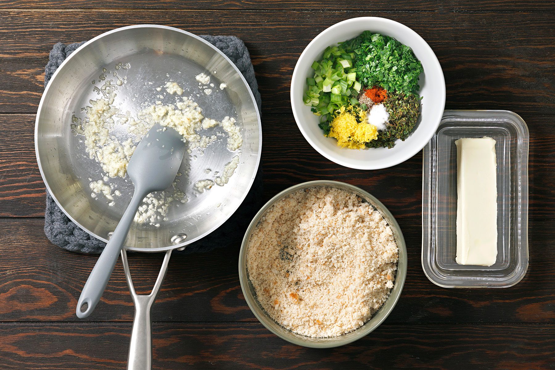 Ingredients for cooking: a pan with sautéed garlic, a bowl of breadcrumbs, a bowl with herbs and spices, and a stick of butter on the side. The items are set on a dark wooden table.