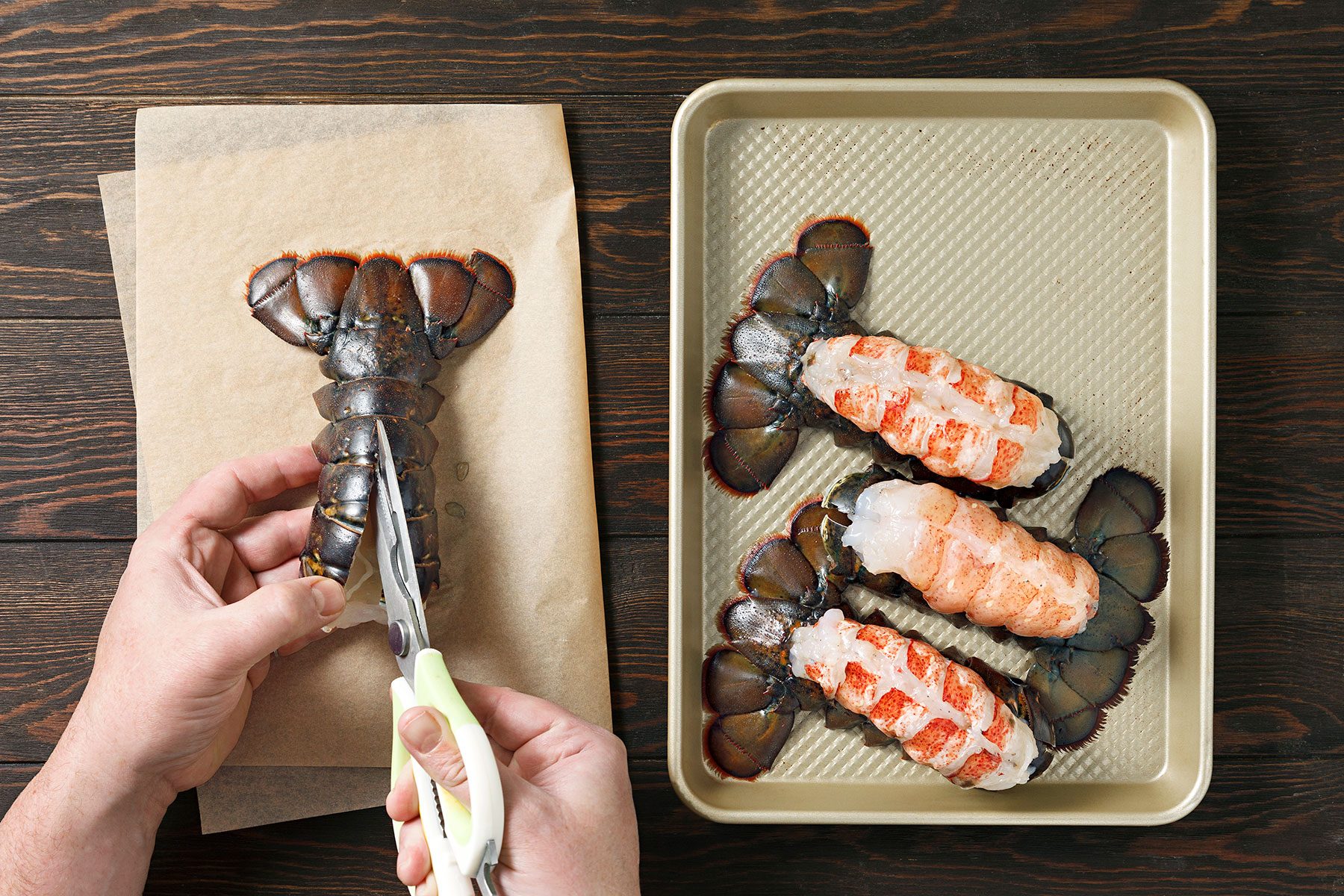 A person uses scissors to cut through the shell of a raw lobster tail on parchment paper. Next to them is a baking sheet with four lobster tails, some split open to reveal the meat.