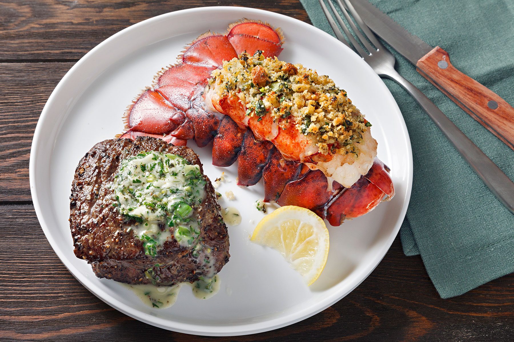 A white plate with a seasoned steak topped with herb butter and a baked lobster tail garnished with breadcrumbs. A lemon wedge is on the side, and silverware rests on a green napkin.