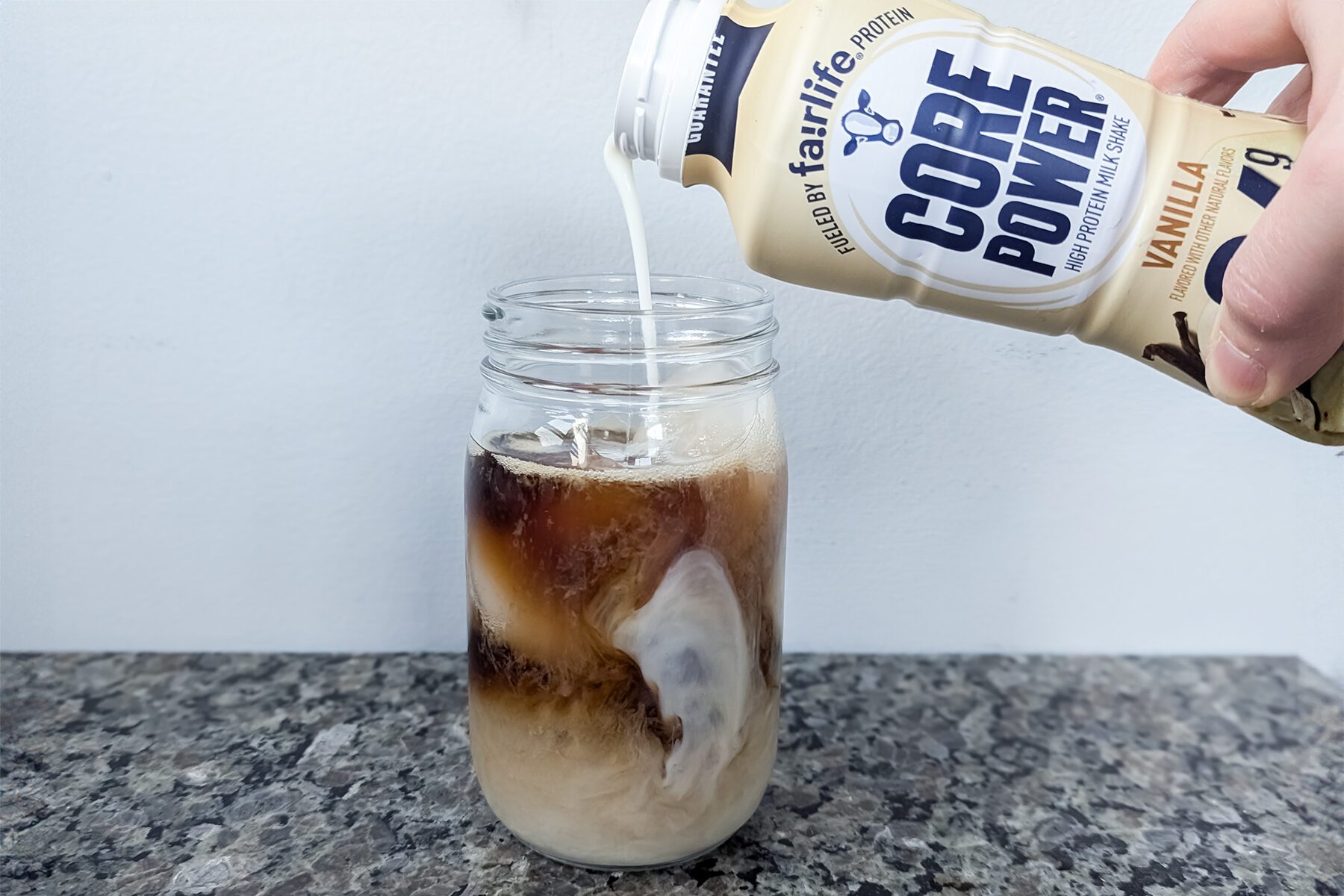 A hand pours Core Power vanilla protein shake into a mason jar filled with iced coffee, creating a swirling effect. The jar is placed on a textured countertop.