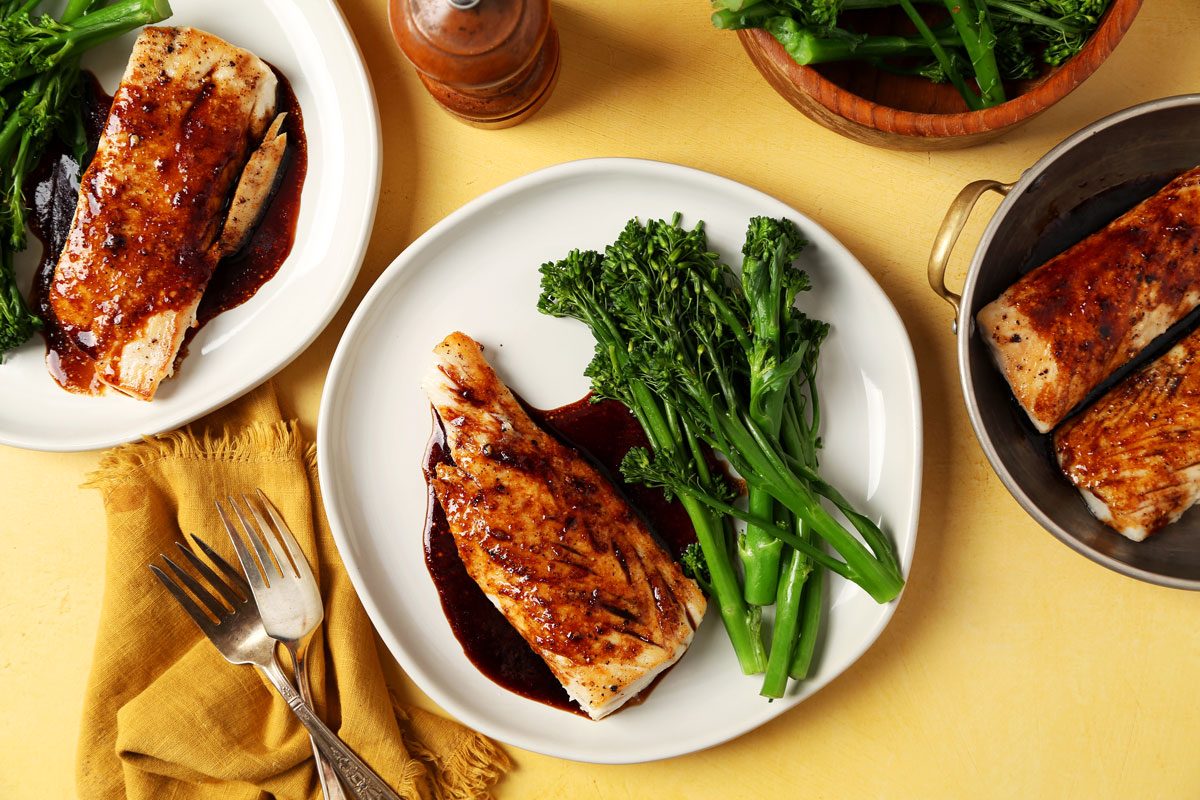 Close up of Taste of Home's Teriyaki Mahi Mahi served on white plates with broccoli rabe on a bright yellow surface.