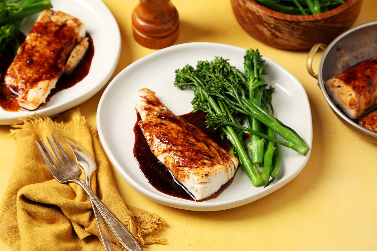 Close up of Taste of Home's Teriyaki Mahi Mahi served on white plates with broccoli rabe on a bright yellow surface.