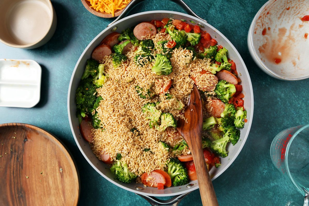 Process of making Taste of Home's Sausage, Rice and Broccoli in a large skillet on a green surface with a green linen.