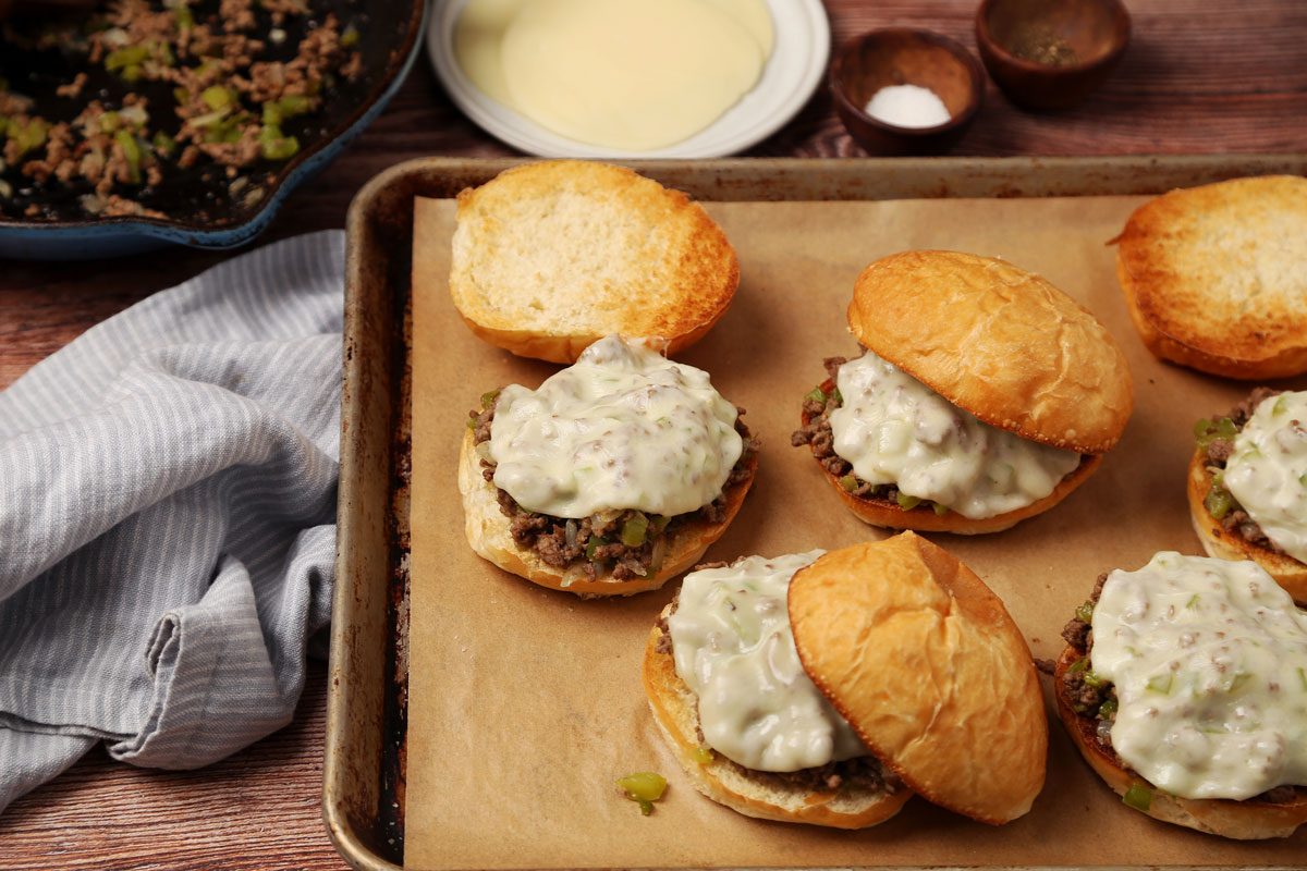 Process of making Taste of Home's Philly Cheesesteak Sloppy Joes on a brown wooden surface. Sandwiches assembled on a baking sheet with melted cheese.