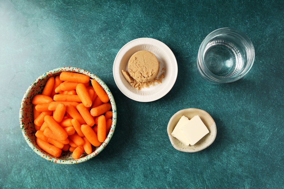 Ingredients for Taste of Home's Glazed Carrots laid out in small bowls on a dark green surface.