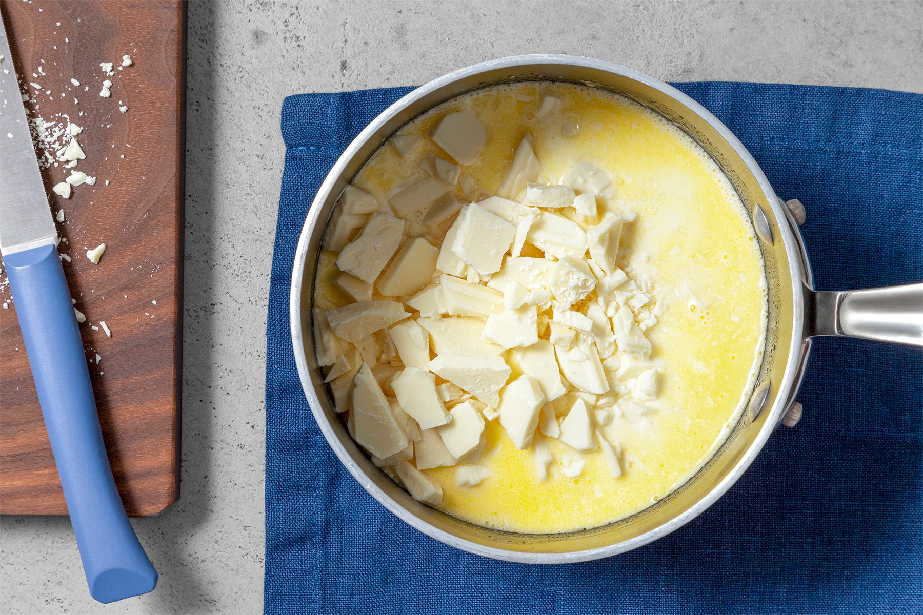 Overhead shot of add white chocolate and remove from the heat; let stand for 5 minutes; blue cloth; knife; wooden board; grey surface;