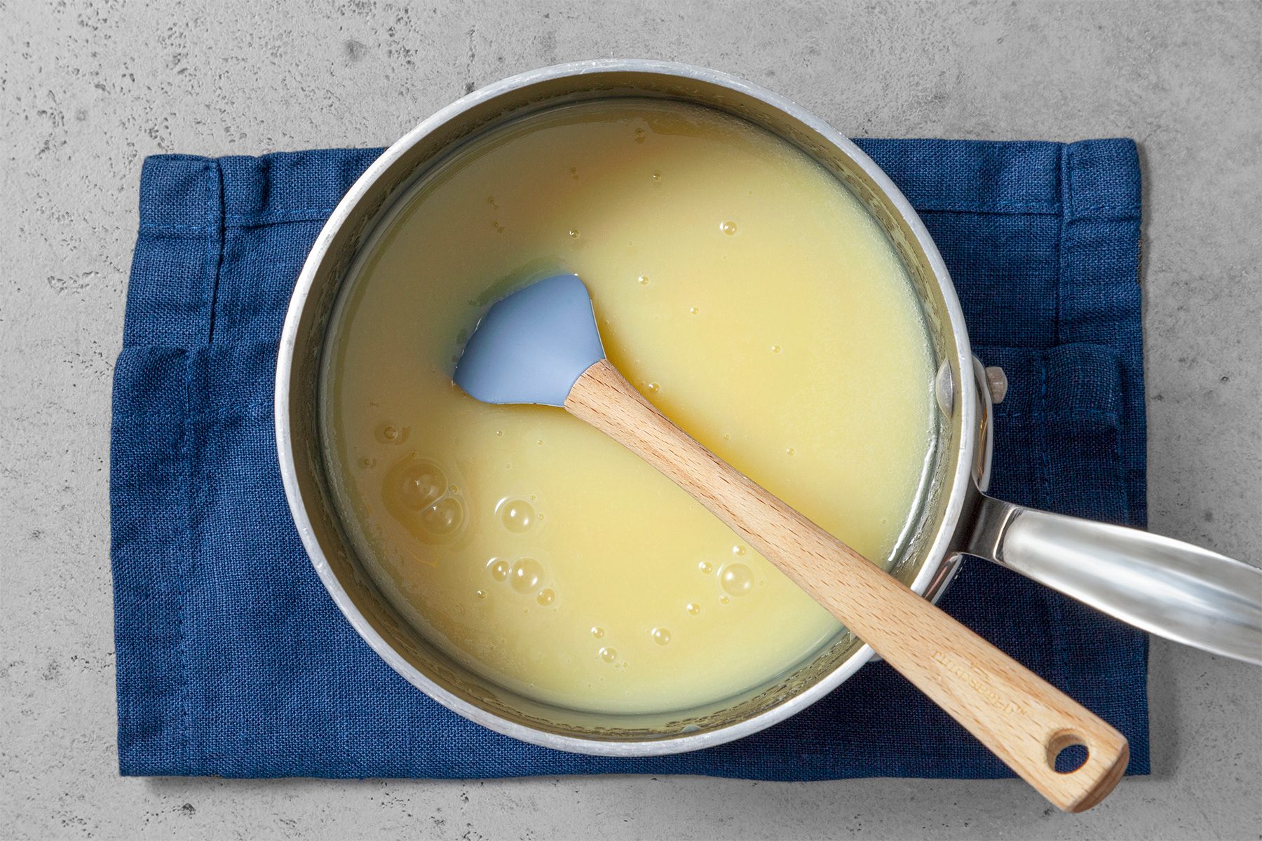 Overhead shot of same saucepan; stir until smooth; spatula; blue cloth; grey surface;
