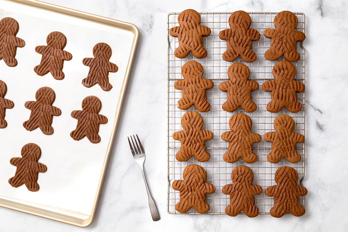 Gingerbread dough cut outs with pressed fork designs alongside a wire rack with baked gingerbread cookies for step five of Wookie Cookie recipe for Taste of Home