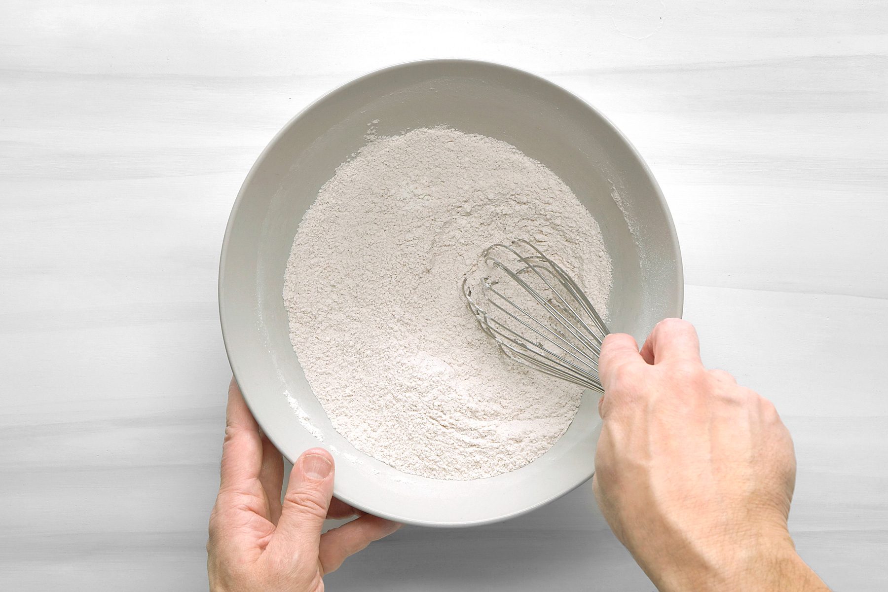 overhead shot of a person's hands holding a bowl with flour and a whisk; the bowl is gray and there is white flour in the bowl; the hands are holding the bowl and whisk; the background is a white surface