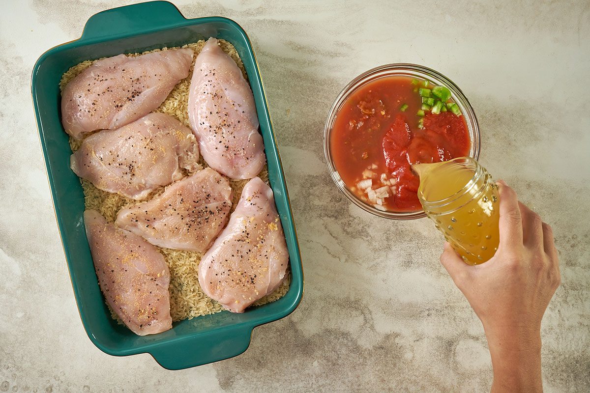 Overhead view of the chicken resting on rice in the baking dish, with bowls of chicken broth, picante sauce, tomato sauce, chopped onion, and green pepper next to the pan, ready to be combined and poured over the chicken for the Taste of Home Arroz con Pollo recipe.