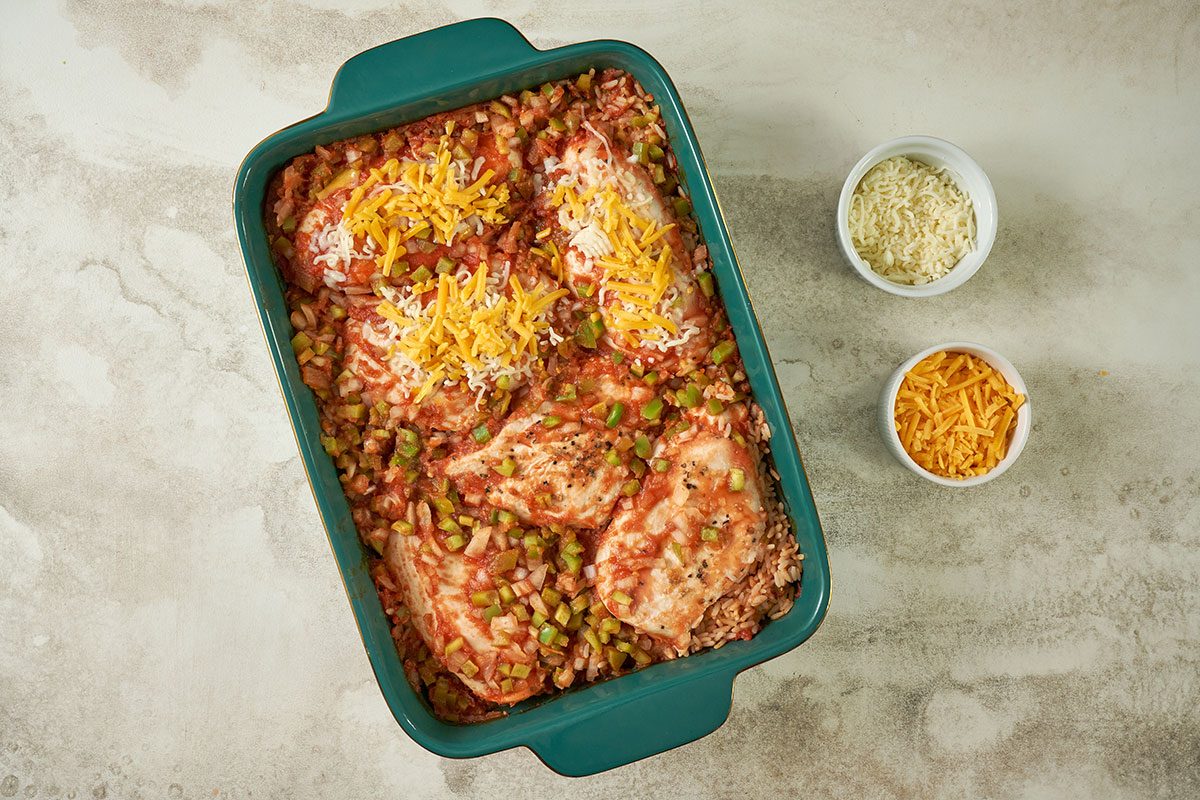 Overhead view of the cheese being sprinkled over the baked chicken and rice mixture, ready to bake uncovered for the Taste of Home Arroz con Pollo recipe.