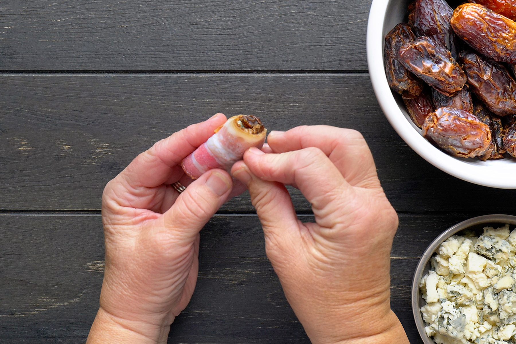 Hands wrapping a date with bacon on a dark wooden surface. A bowl of dates and a dish of crumbled blue cheese are nearby.