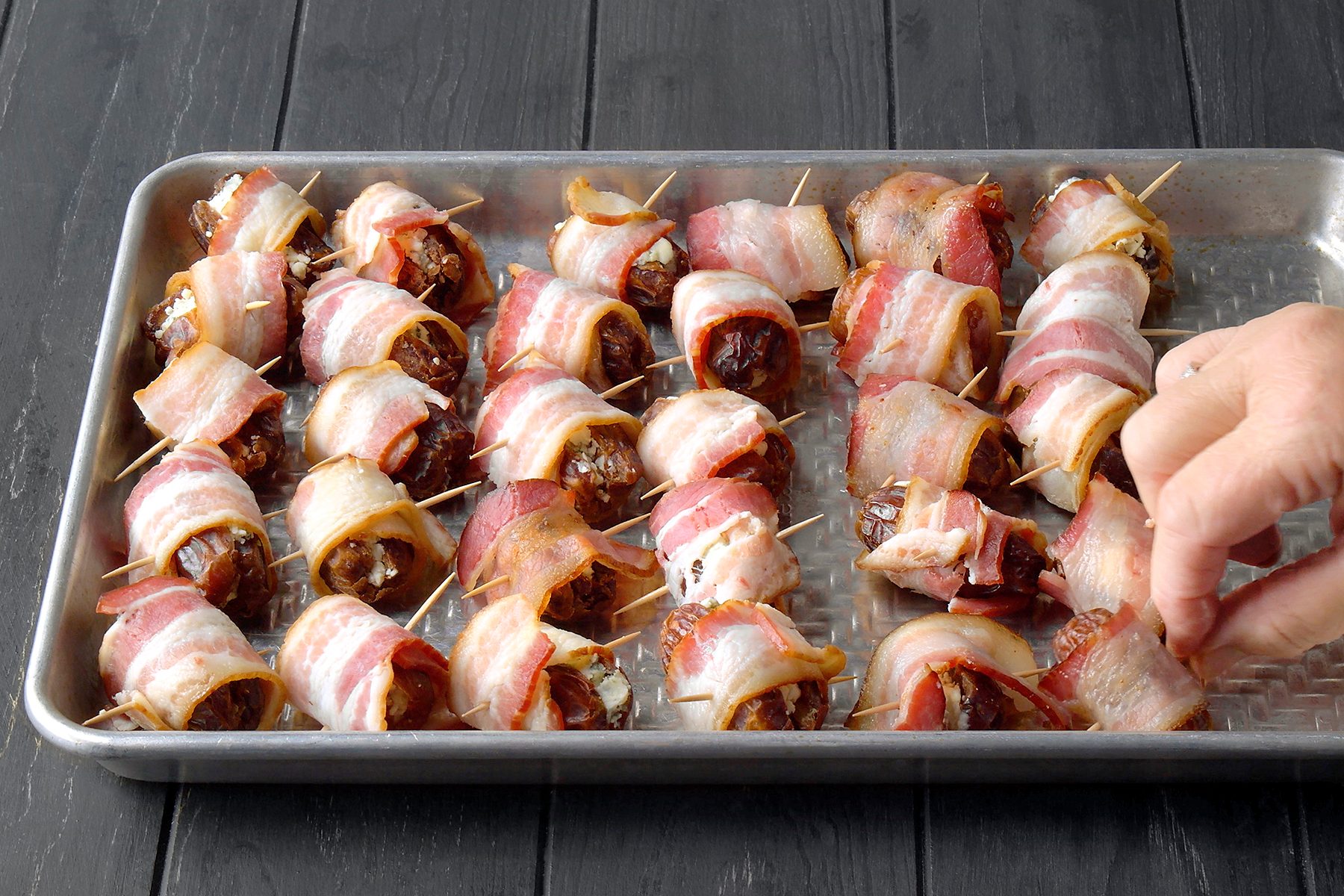 A hand reaches for bacon-wrapped dates on a metal baking tray. The dates are secured with toothpicks and arranged in rows on the tray, which is placed on a dark wooden surface.