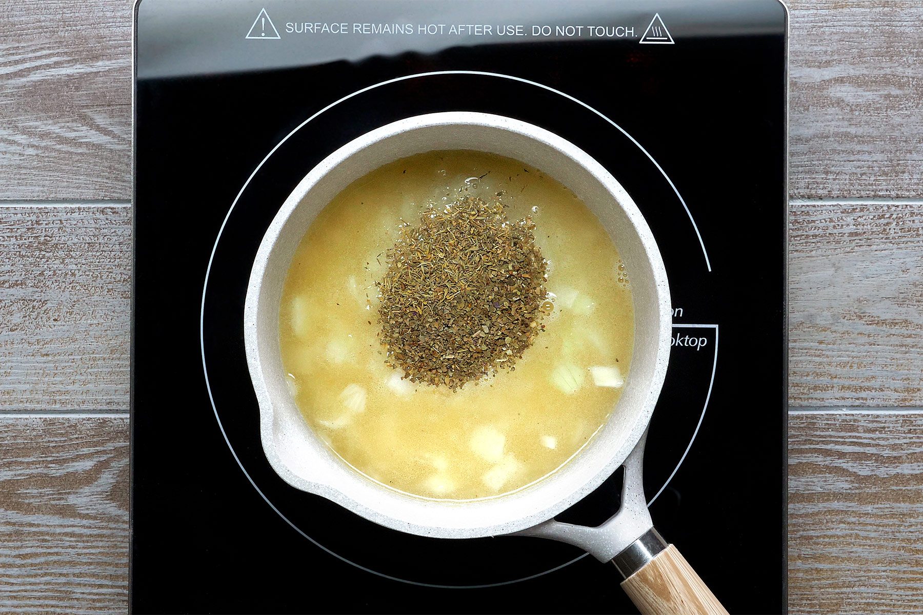 overhead shot of a white pot with a wooden handle sitting on a black electric stovetop; there are some herbs and chopped onions in the pot; the pot appears to be sitting on the hot burner