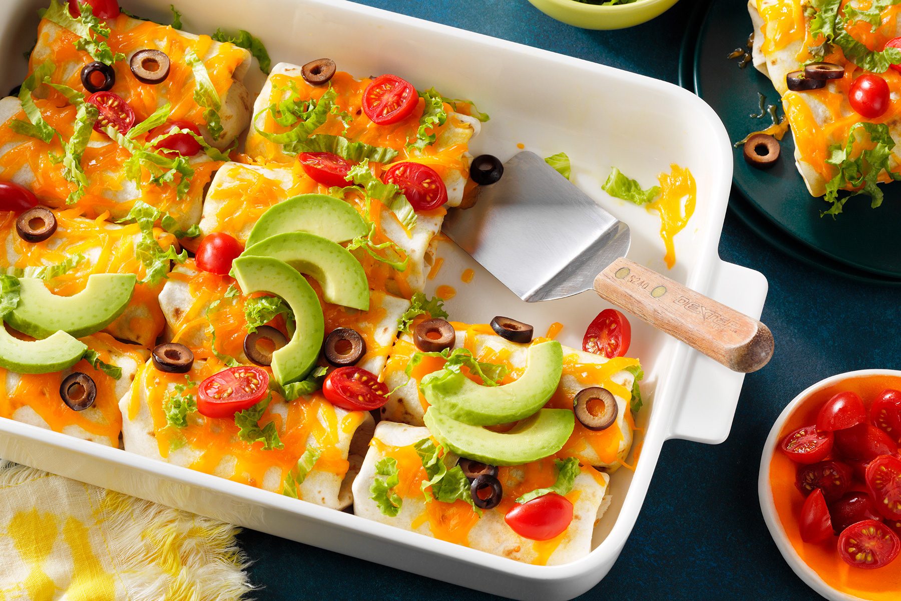 A white baking dish filled with enchiladas topped with melted cheese, avocado slices, cherry tomato halves, and black olive slices. A spatula is resting on the edge. A small bowl of cherry tomatoes is nearby.