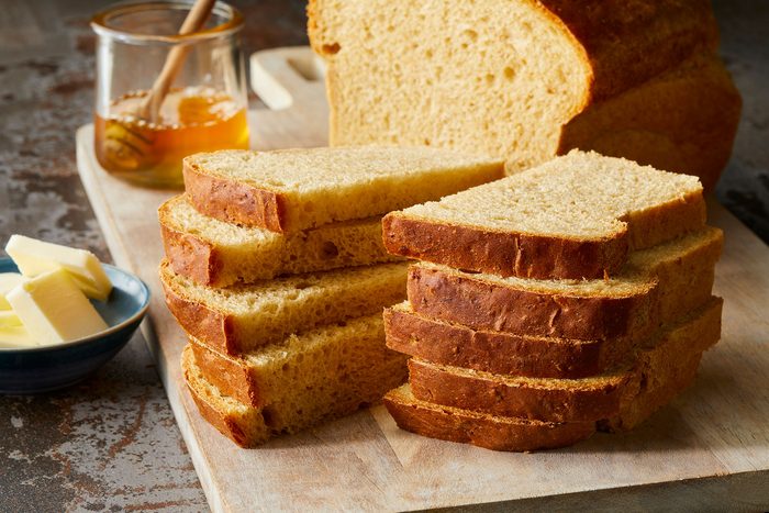 Slices of brown bread are neatly stacked on a wooden cutting board. A jar of honey with a dipper sits nearby, along with a blue bowl of butter. A loaf of unsliced bread is in the background.