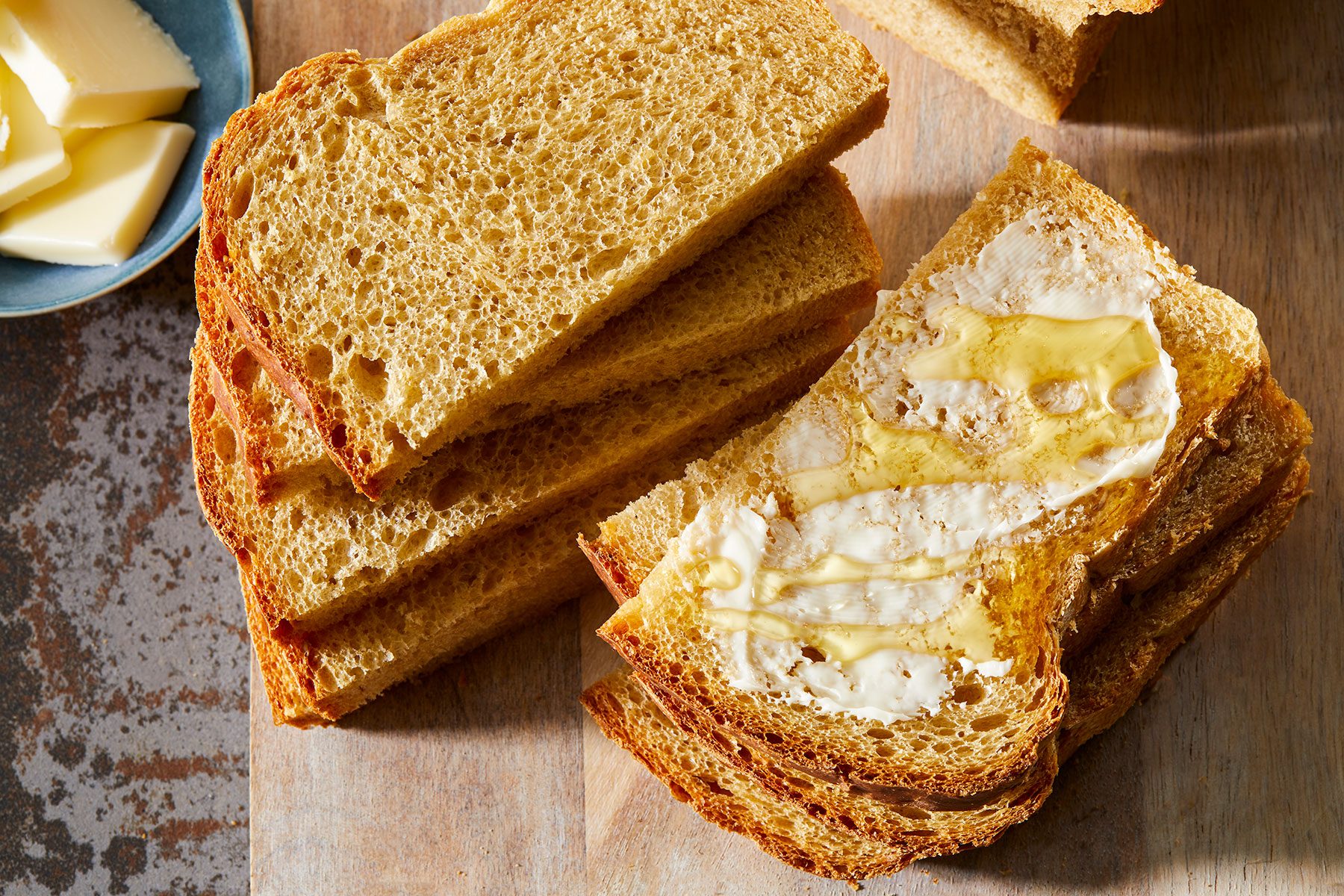Slices of brown bread on a wooden surface, with one slice partially spread with butter. A small plate with butter pieces is visible in the corner.