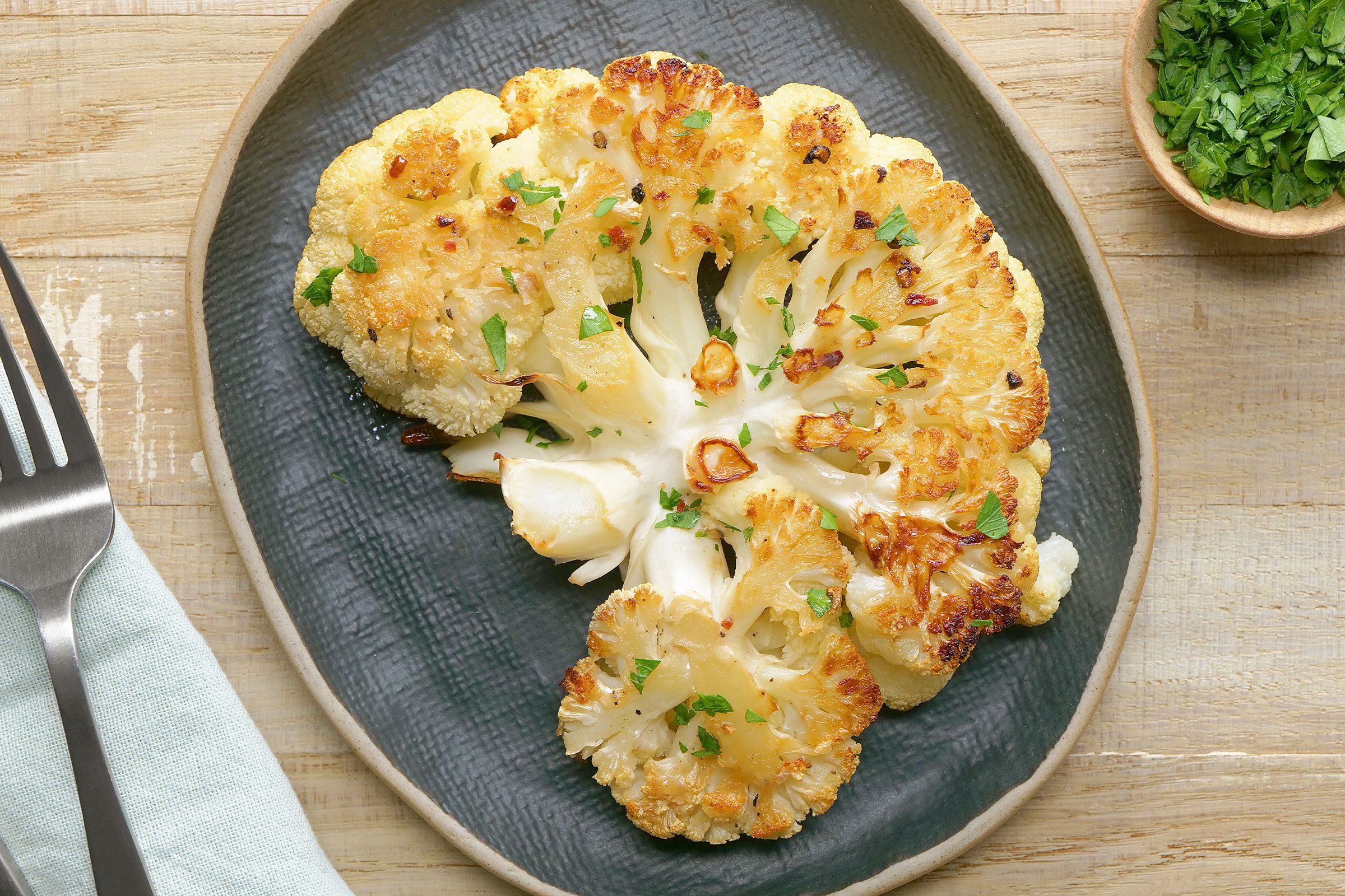 Cauliflower Steak sprinkled with parsley placed on a grey dish with a fork on side