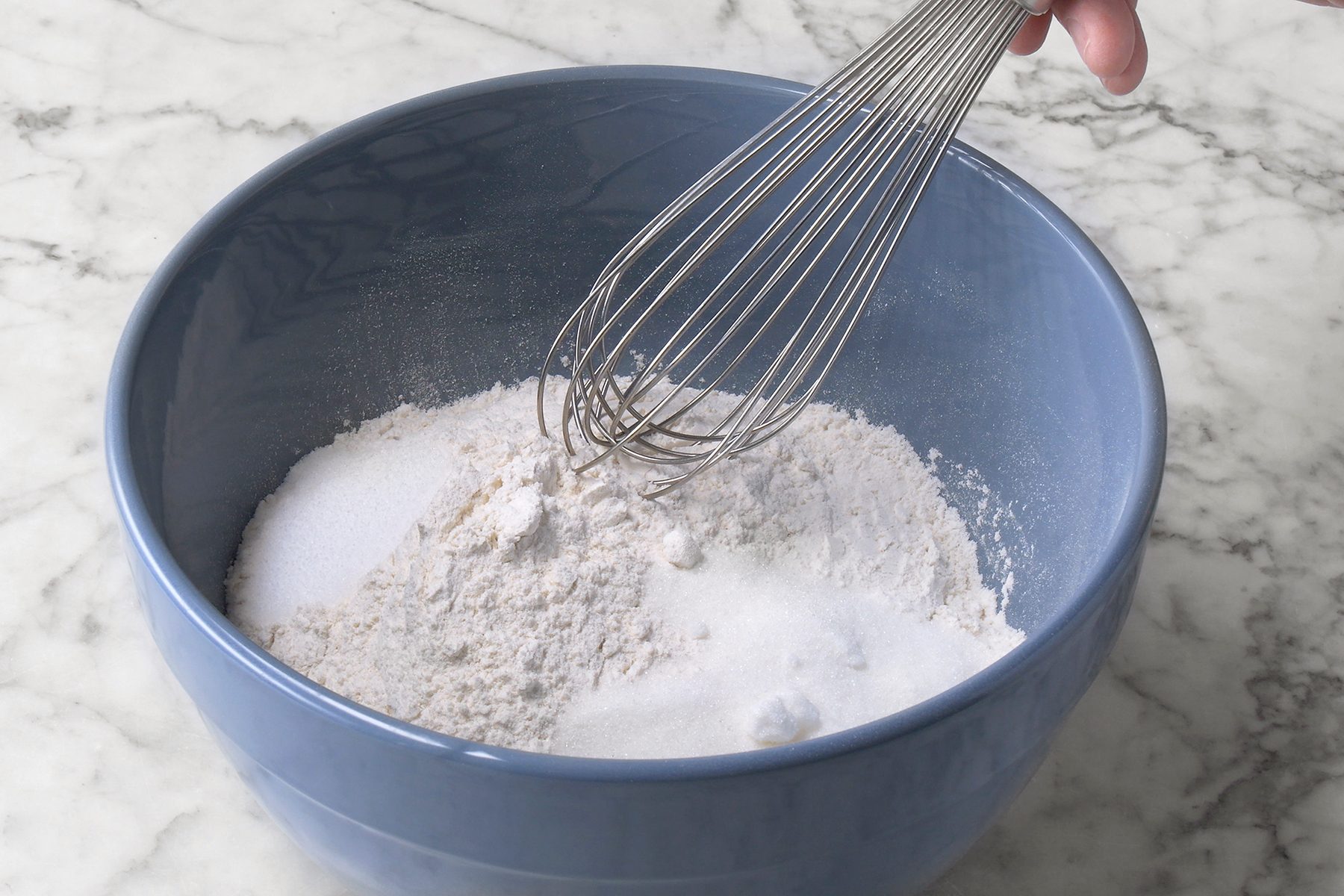 A hand holding a metal whisk is poised over a blue bowl filled with flour. The bowl is set on a marble countertop.