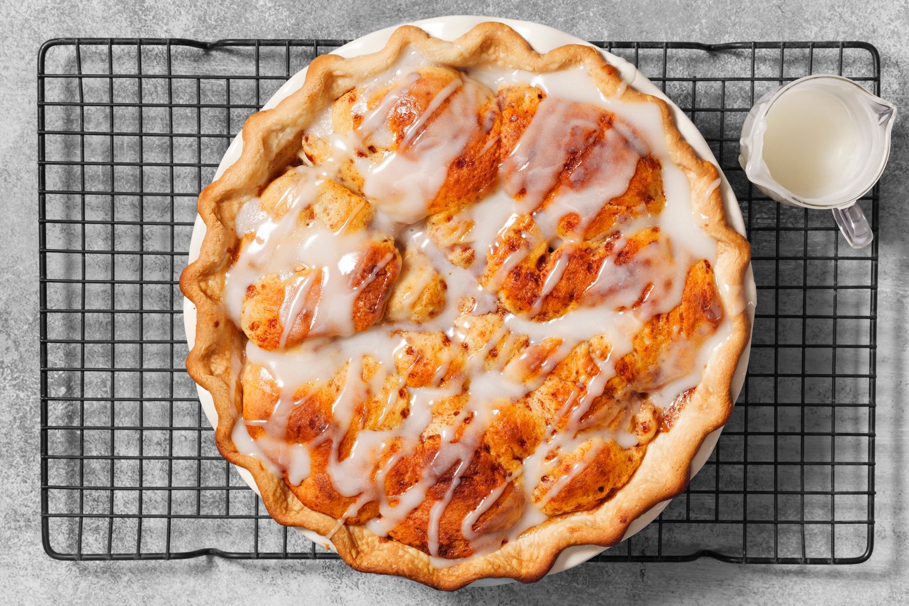 overhead shot of a baked dessert, likely a pie, placed on a black wire cooling rack, featuring a golden-brown crust with fluted edges, a filling resembling cinnamon rolls or baked dough pieces, and a drizzle of white icing over the top, a small pitcher containing extra icing placed to the side