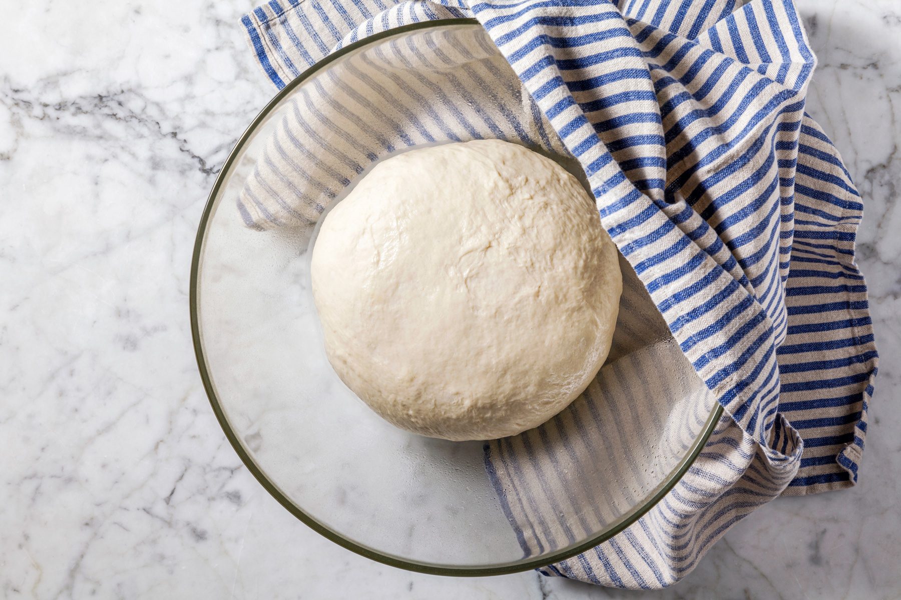 Overhead shot of placing dough in a greased bowl, turning to grease the top; covering and letting rise for 1 hour until doubled, on a marble surface with a napkin.