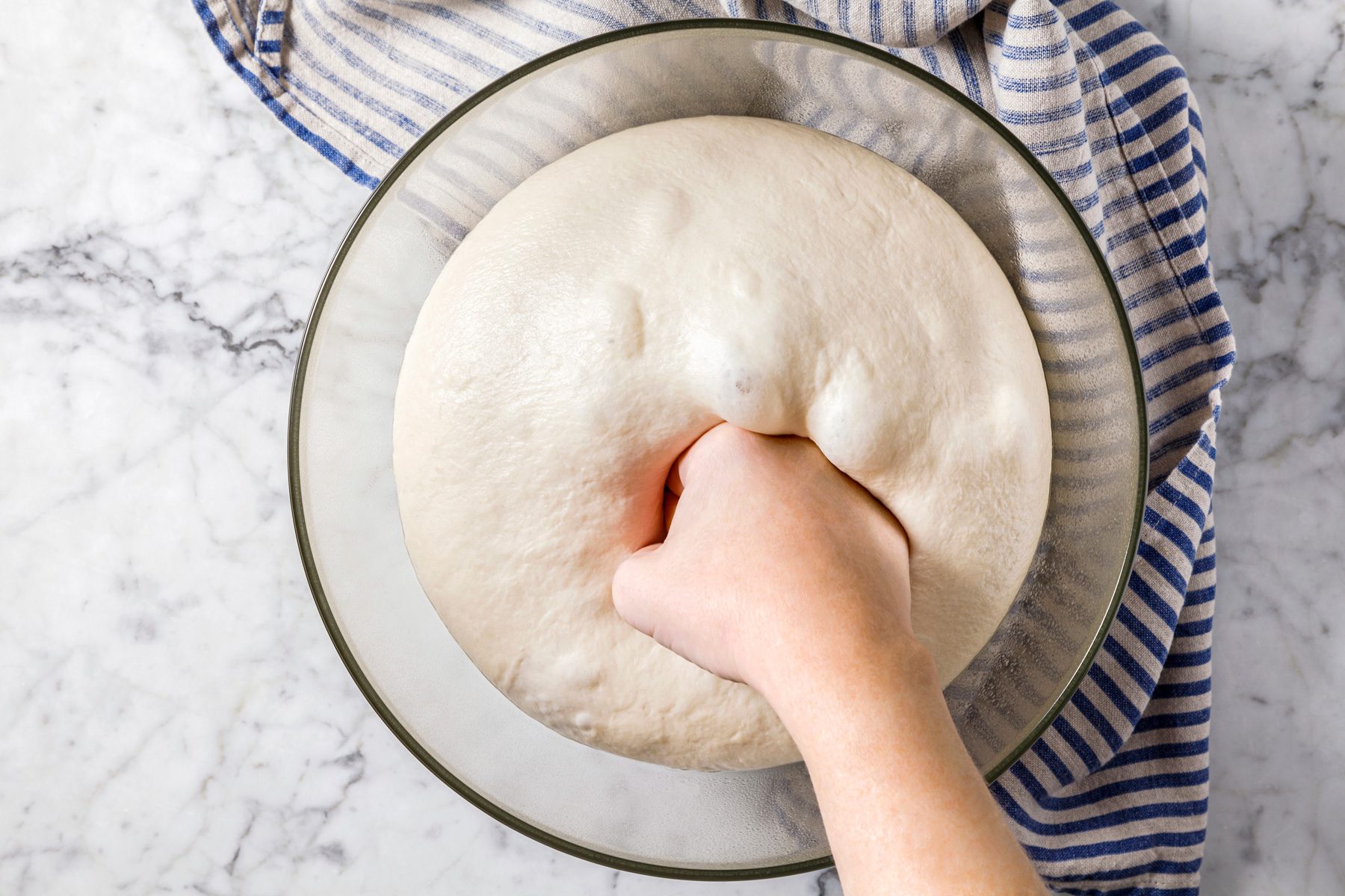 Overhead shot of punching down dough on a marble surface, with a napkin nearby.