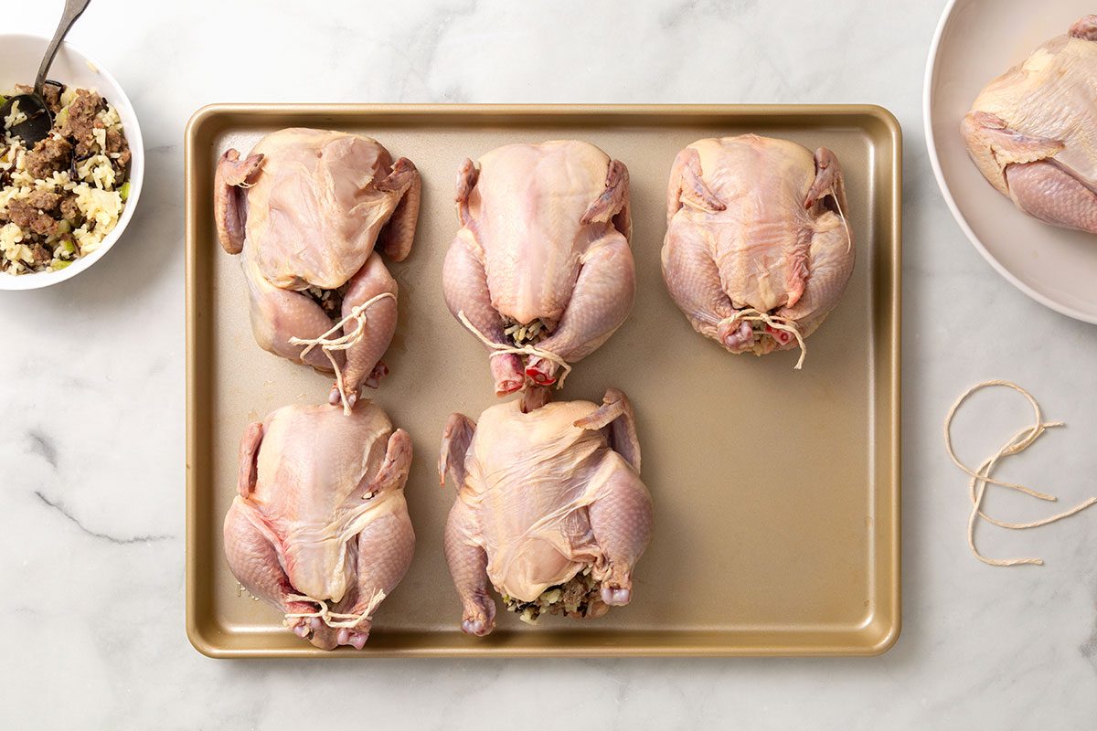 Cornish hens stuffed with rice stuffing mixture on a baking pan. Each with legs tied together with twine for step three of Cornish Game Hens with Wild Rice Stuffing recipe for Taste of Home