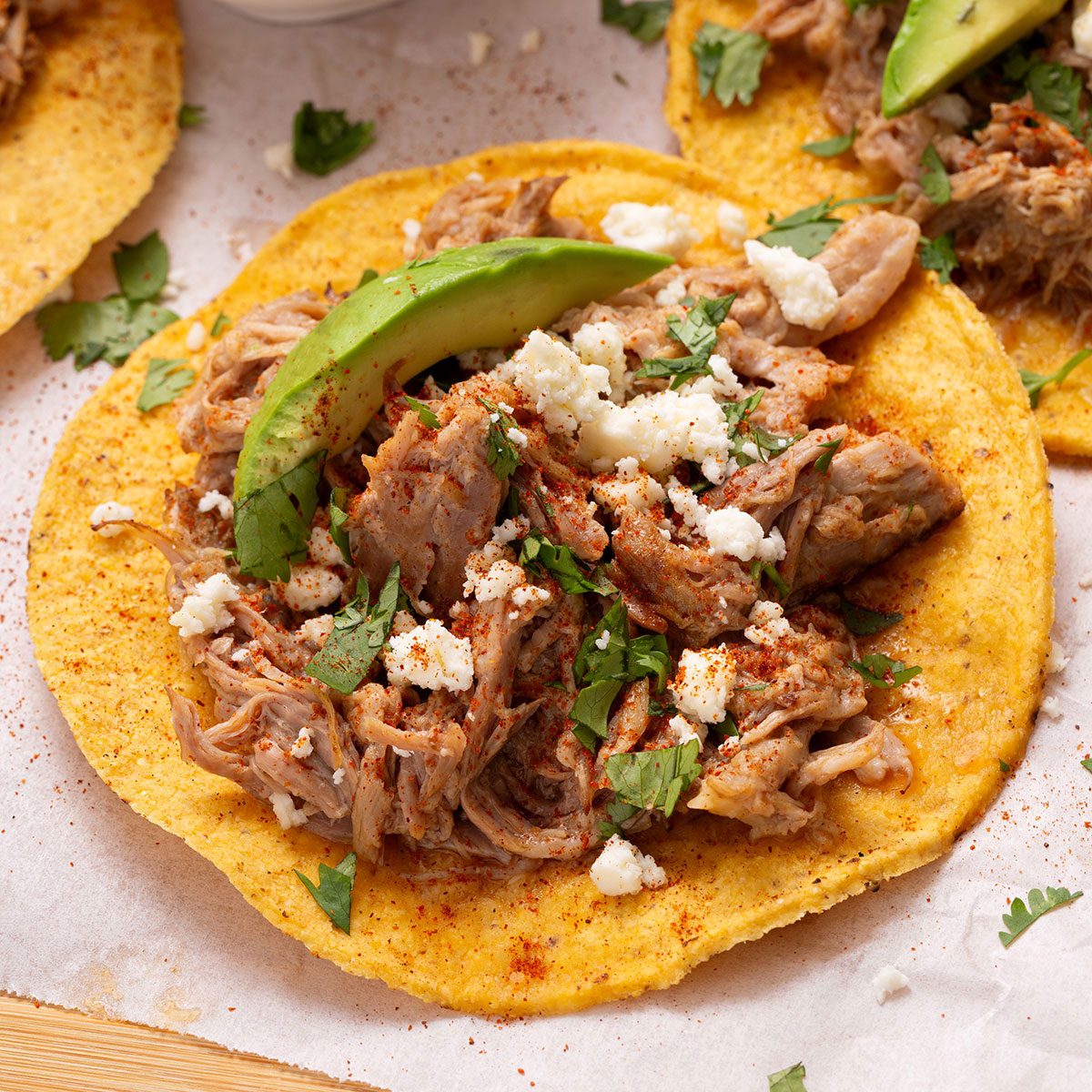 Tostada shell topped with chipotle shredded pork, chopped cilantro, crumbled queso fresco and avocado slices on a wooden cutting board for Taste of Home