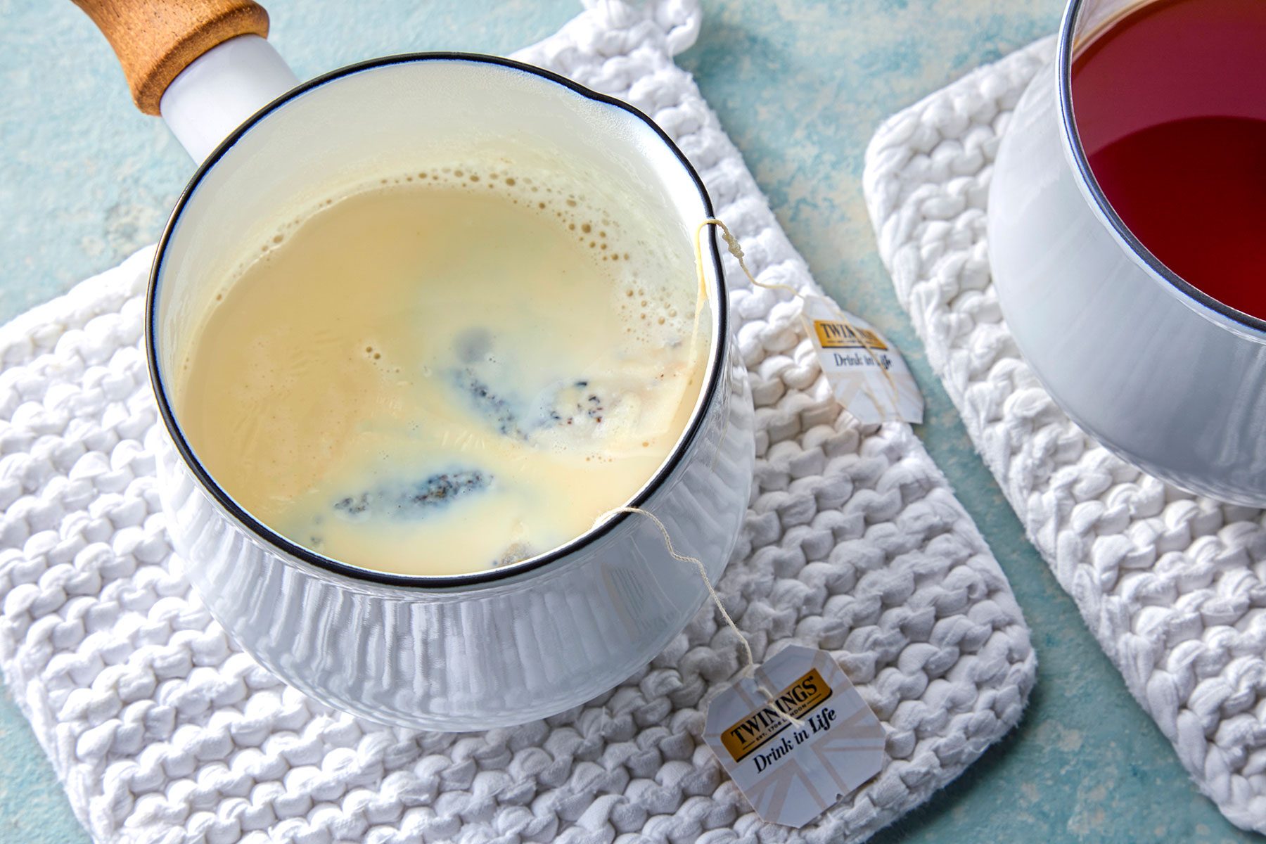 A white saucepan containing creamy milk steeped with two tea bags rests on a white knitted mat. Another cup with a deep red liquid sits nearby on a similar mat. The tea bag tags are visible, labeled "TWININGS Drink In Life.