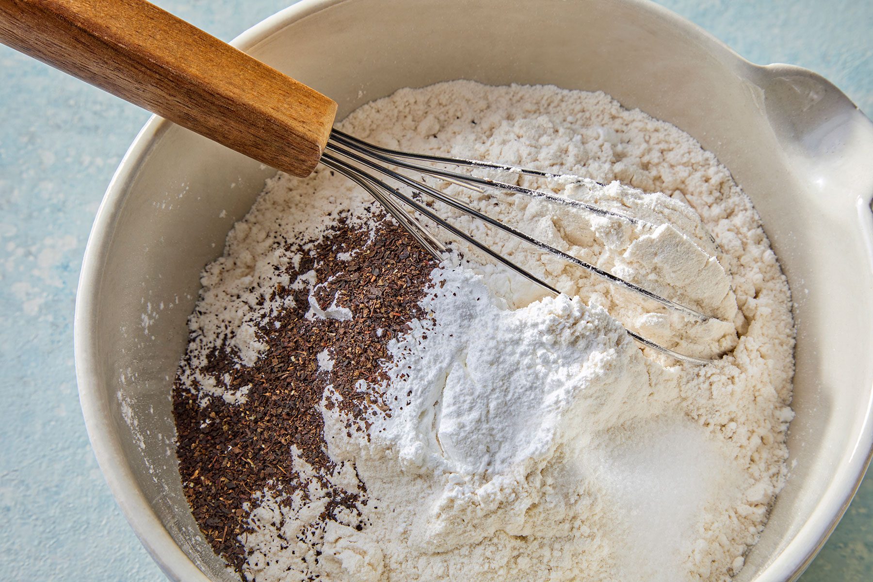 A mixing bowl containing flour, seeds, and spices, with a whisk resting on top.