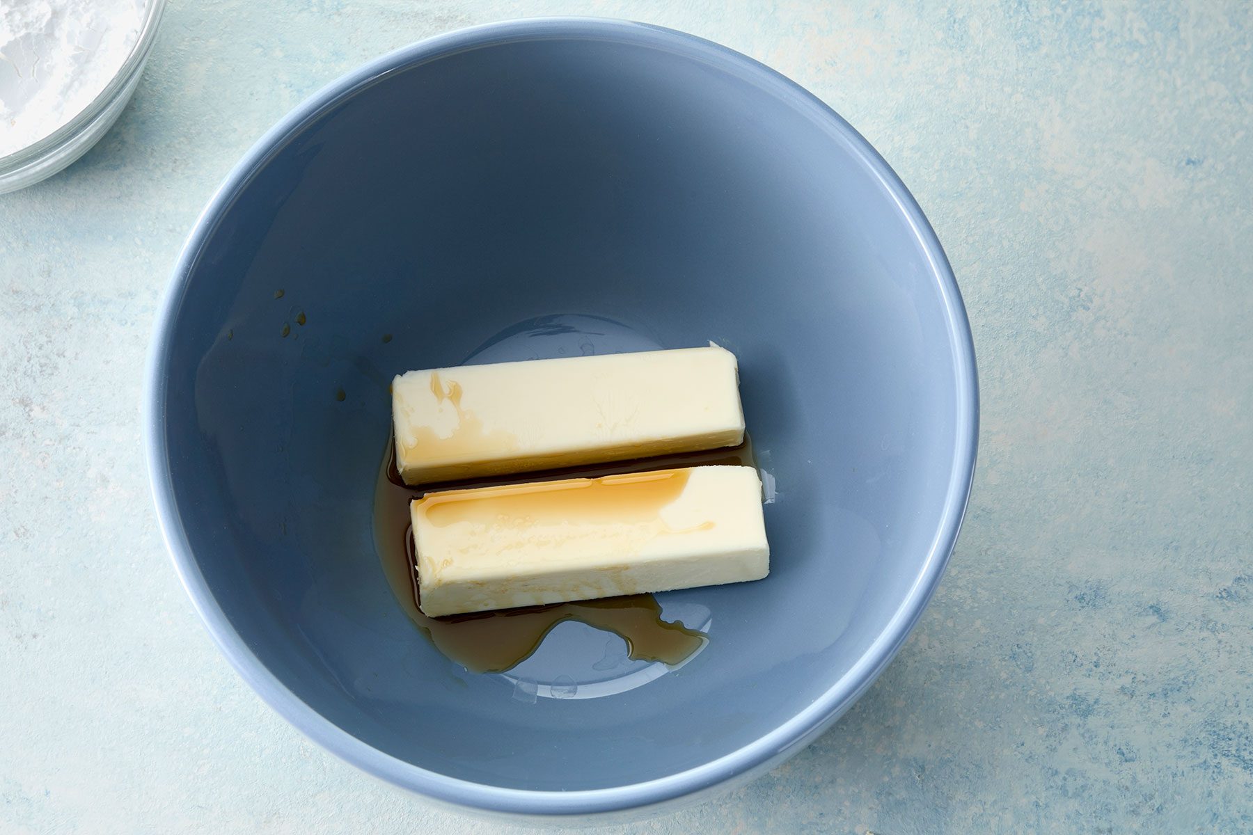 A blue bowl containing two sticks of butter with some liquid, possibly vanilla extract, drizzled over them. The bowl is placed on a light blue textured surface.