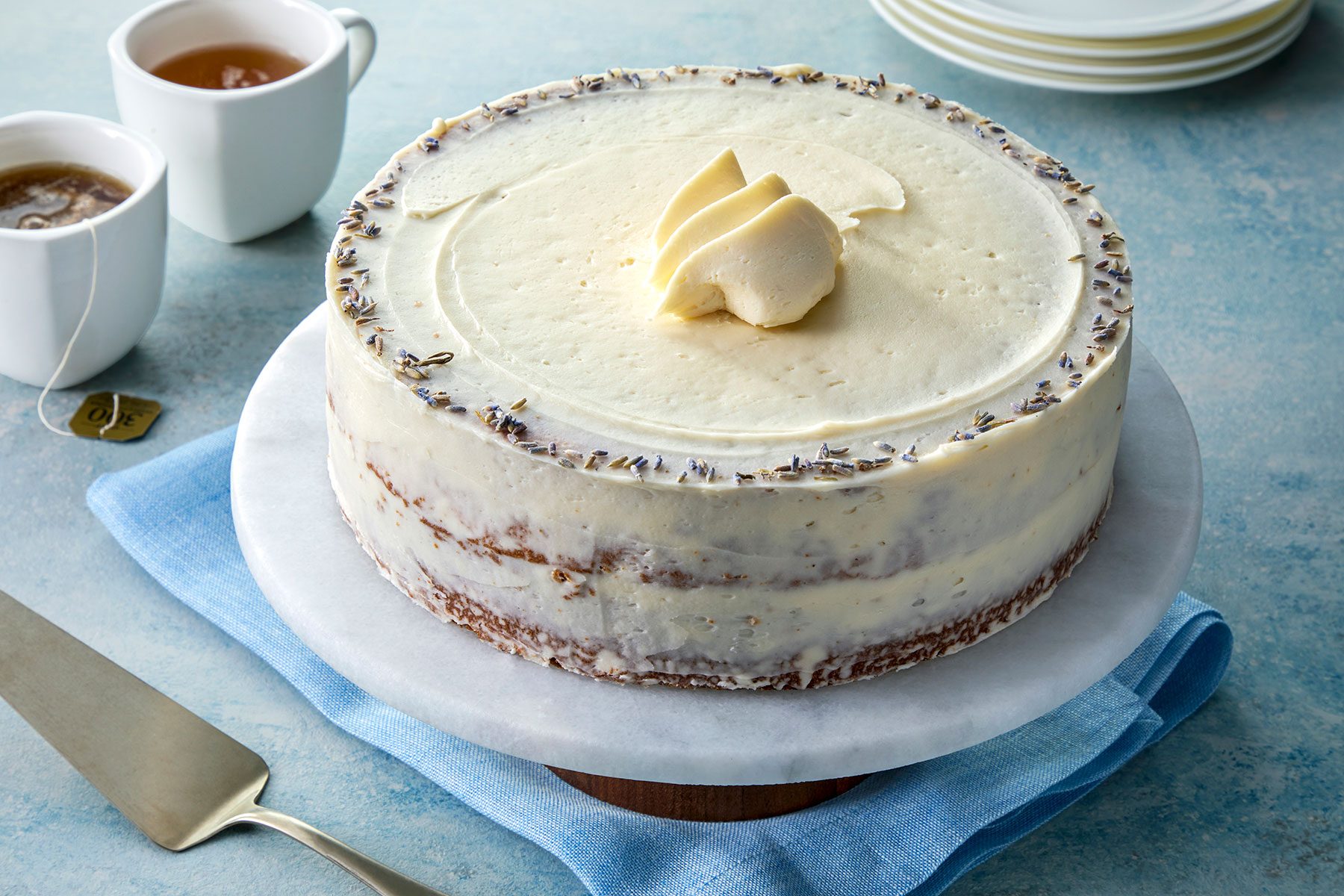 A frosted round cake with a light beige layer, topped with a decorative swirl of butter or cream and sprinkled with small purple flowers. The cake sits on a marble platter placed on a blue cloth, with two cups of tea and a serving utensil nearby.