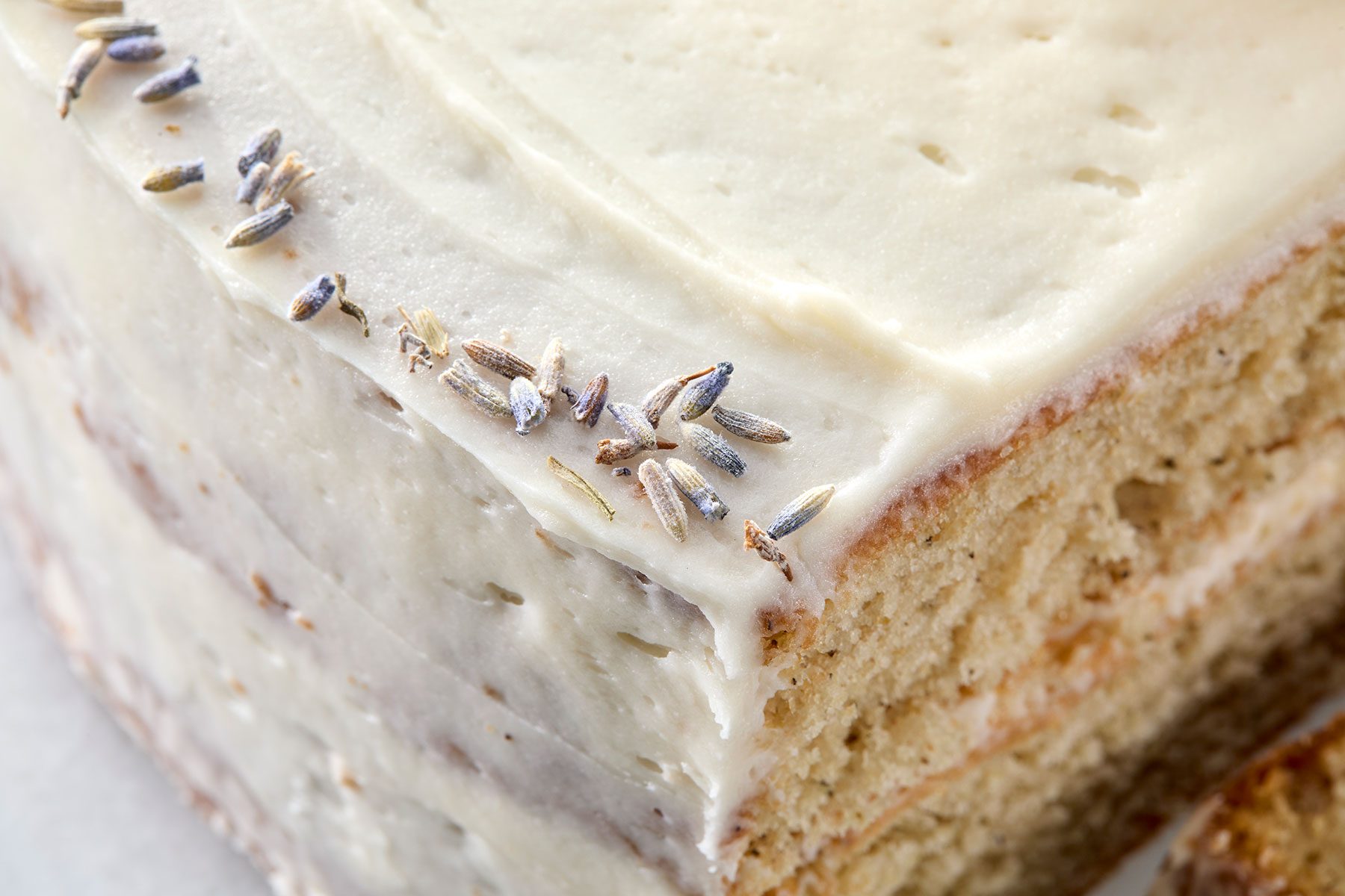Close-up of a slice of cake with creamy white frosting, topped with lavender sprigs. The cake layers are visible and the texture appears soft and moist.