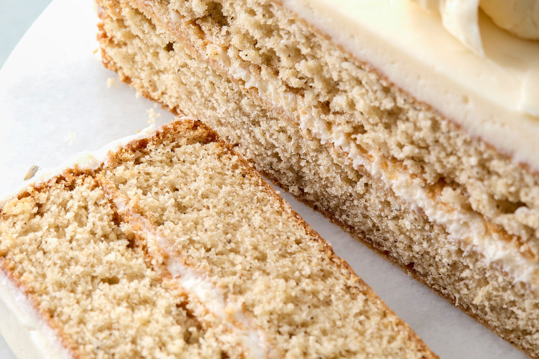 Close-up of a sliced vanilla cake with visible layers of frosting. The texture appears moist and crumbly, with a light cream-colored icing on top. The cake is placed on a white surface.