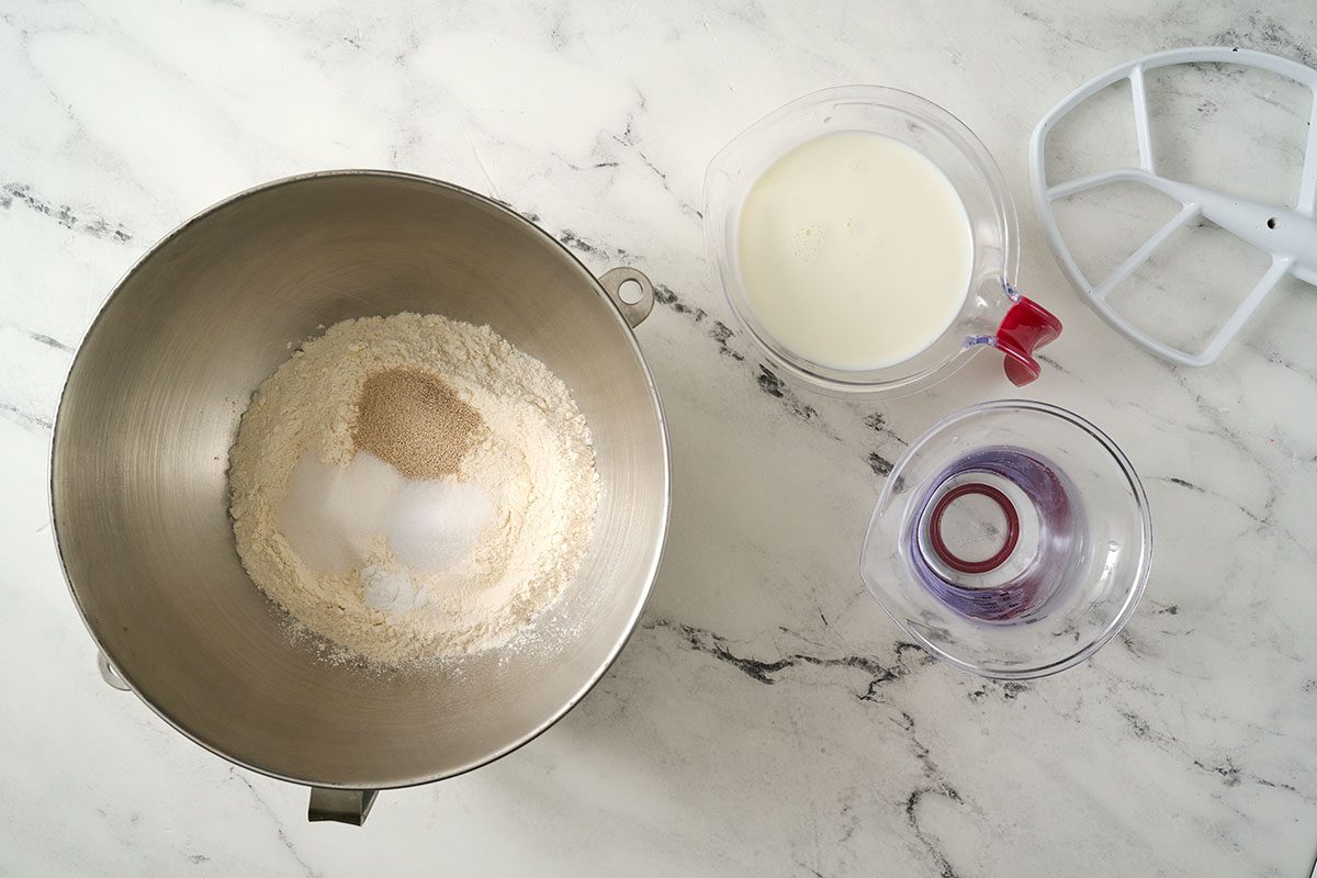 Overhead view of the flour, yeast, sugar, salt, and baking soda being combined with warm milk and water in a large bowl, using a stand mixer on low speed for the Taste of Home English Muffin Bread recipe.