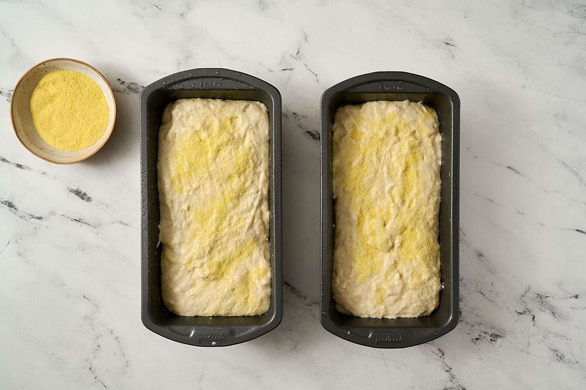 Overhead view of the stiff batter spread in two greased loaf pans, sprinkled with cornmeal on top, and covered to rise for the Taste of Home English Muffin Bread recipe.