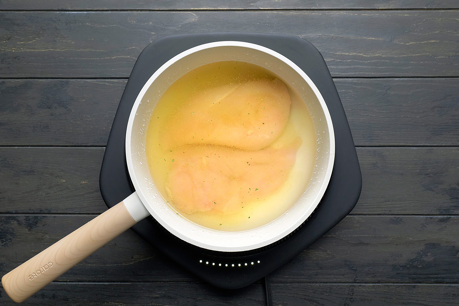 overhead shot of a white pan with a wooden handle on a black induction cooktop; the pan is filled with broth and two chicken breasts; the cooktop is on a black wooden surface