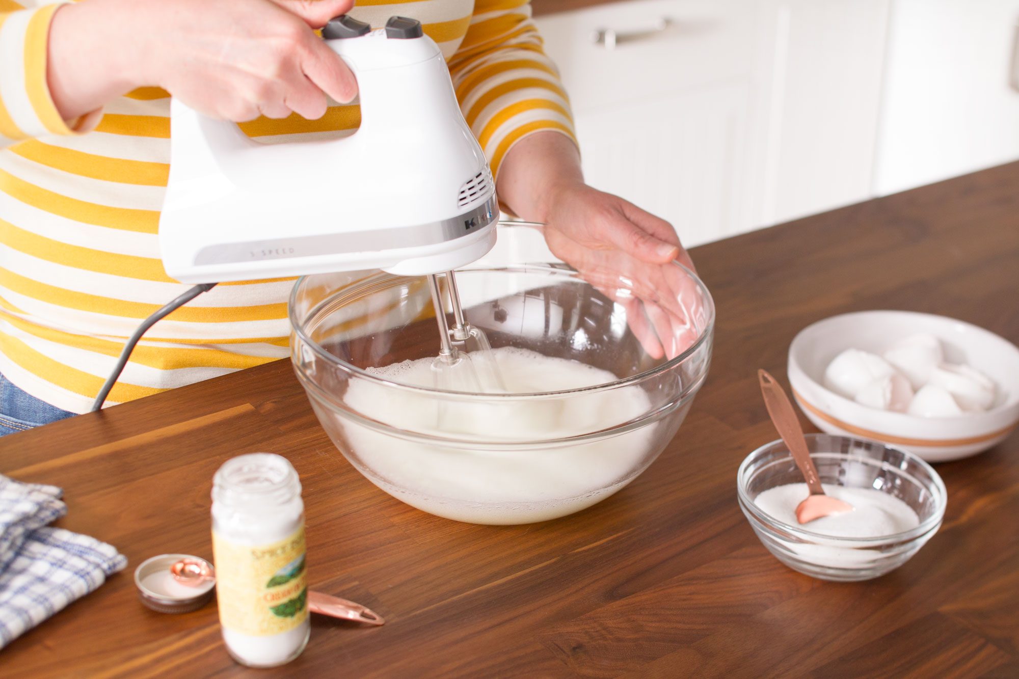 A person wearing a yellow and white striped shirt is using a white electric hand mixer to beat ingredients in a large glass bowl. There are eggs, a jar, a folded cloth, and measuring spoons on the wooden countertop.
