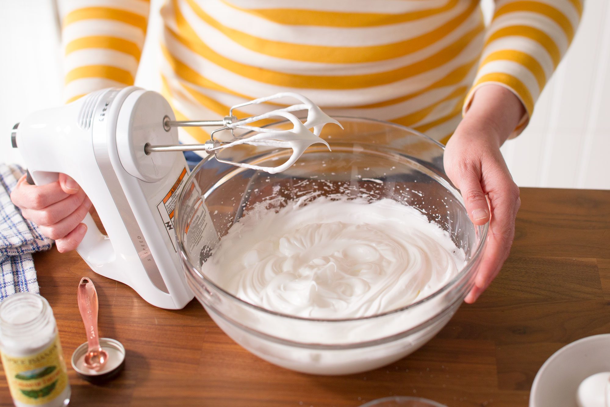 A person in a yellow and white striped shirt uses a hand mixer to whip cream in a glass bowl on a wooden countertop.