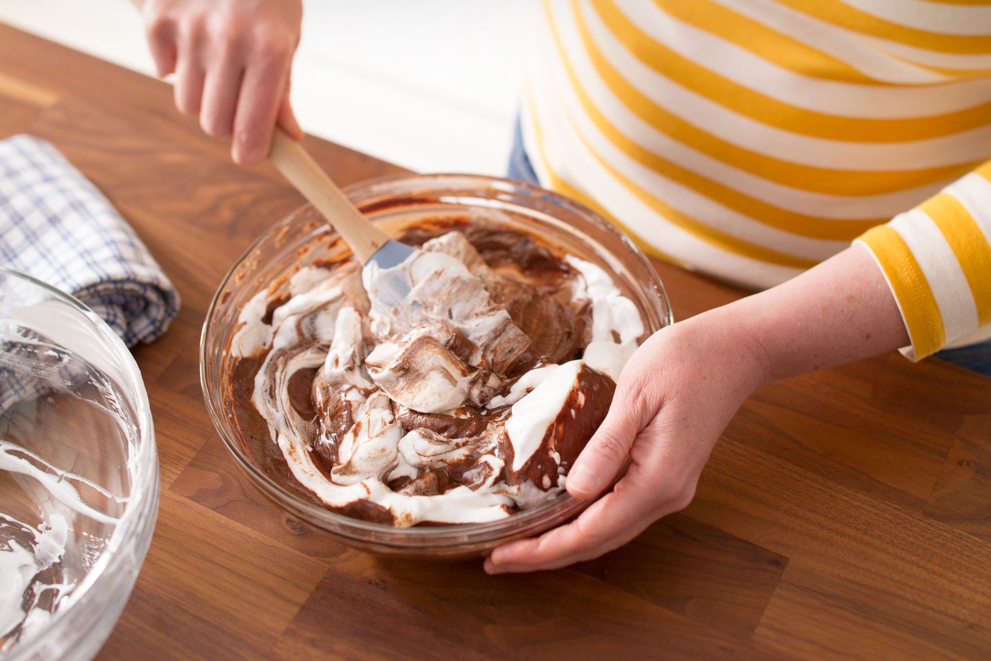 A person in a yellow and white striped shirt is mixing chocolate batter and white cream in a glass bowl with a spatula on a wooden countertop.